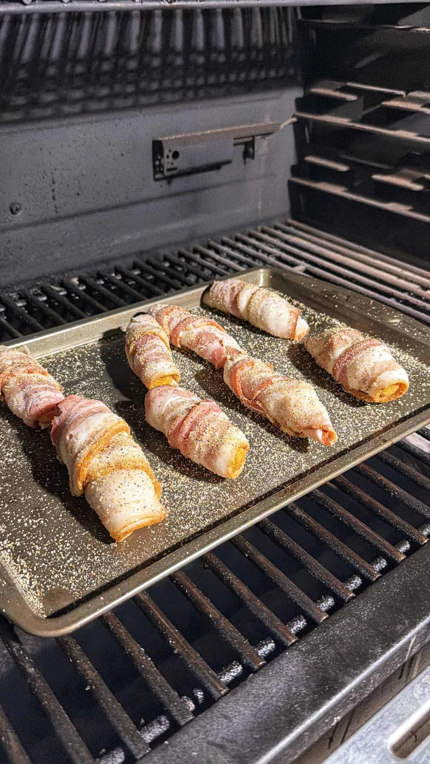 Bacon-wrapped stuffed jalapeños on a baking tray inside a grill, seasoned with pepper and ready to be cooked.