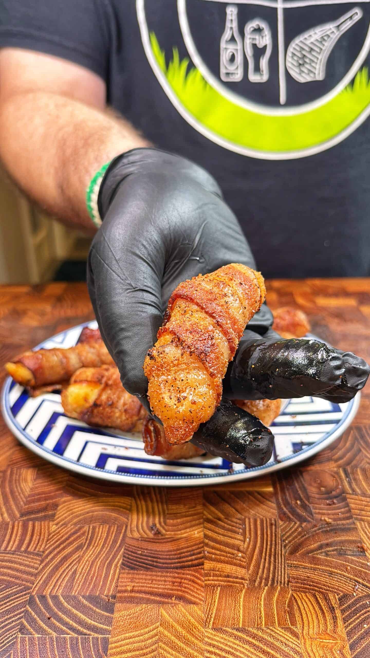 A person wearing a black glove holds a piece of bacon-wrapped food, with more pieces on a blue and white plate in the background, all on a wooden cutting board.