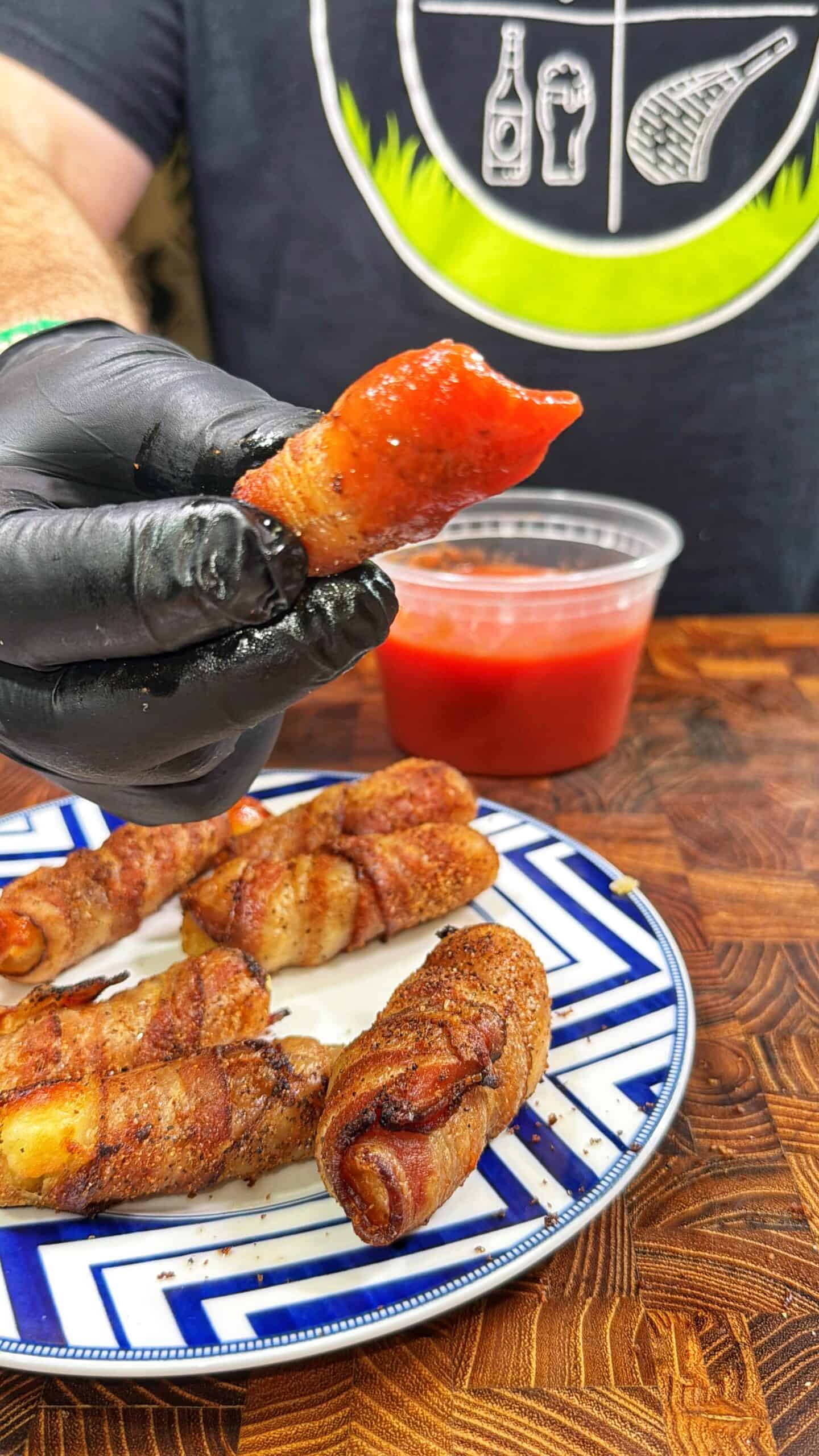 A person wearing black gloves holds up a bacon-wrapped snack, dipped in red sauce. More bacon-wrapped snacks are on a blue and white plate, with a container of sauce and a wood surface beneath.
