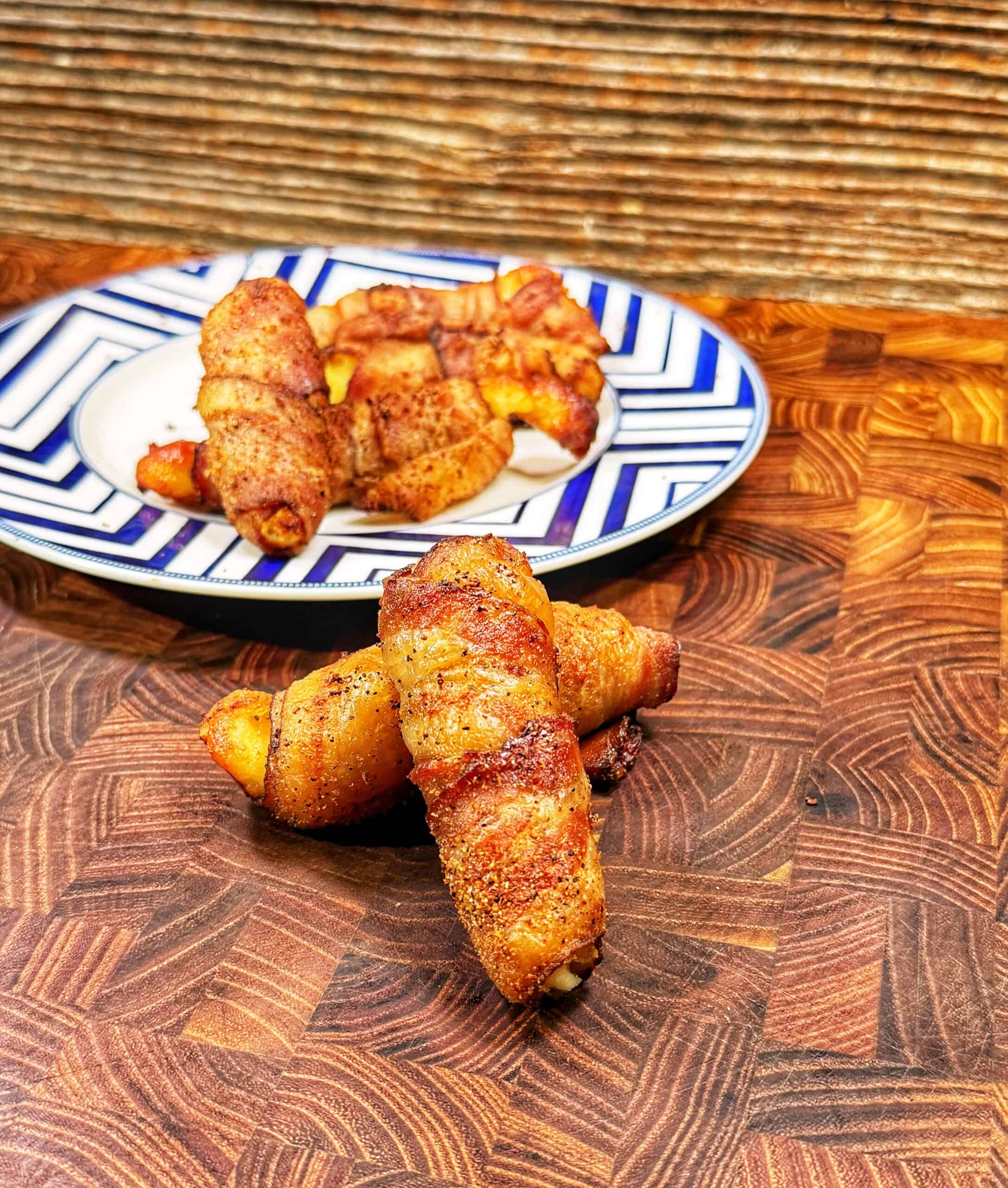 Three bacon-wrapped snacks are stacked on a wooden surface, with more on a blue and white patterned plate in the background. The bacon appears crispy and seasoned.