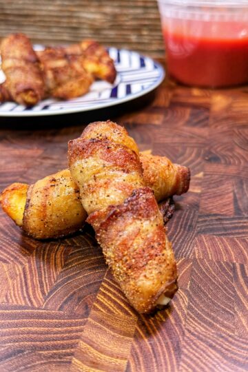 Two pieces of bacon-wrapped food sit on a wooden surface, with more pieces on a plate in the background and a plastic container of red dipping sauce nearby.