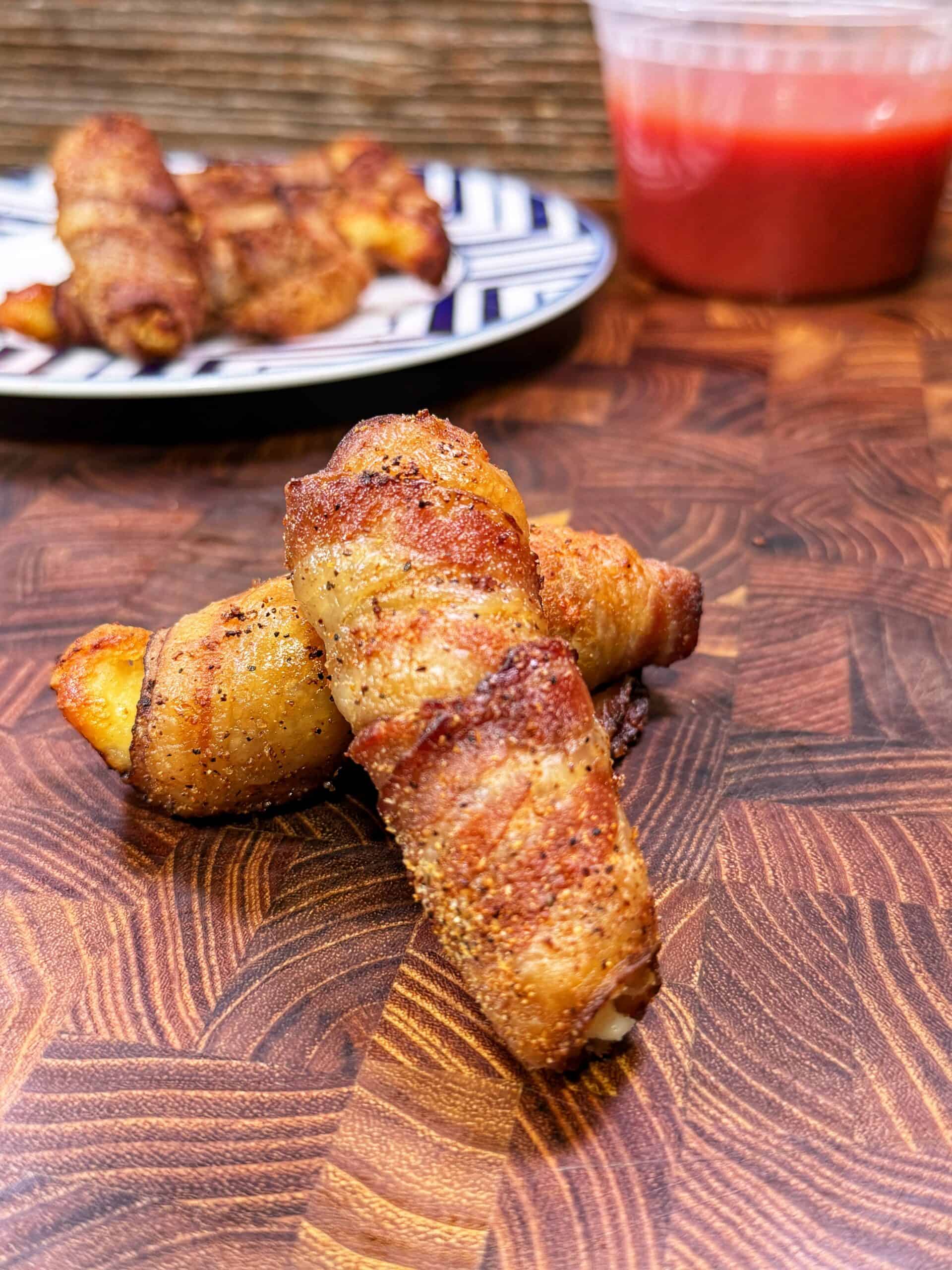 Two pieces of bacon-wrapped food sit on a wooden surface, with more pieces on a plate in the background and a plastic container of red dipping sauce nearby.