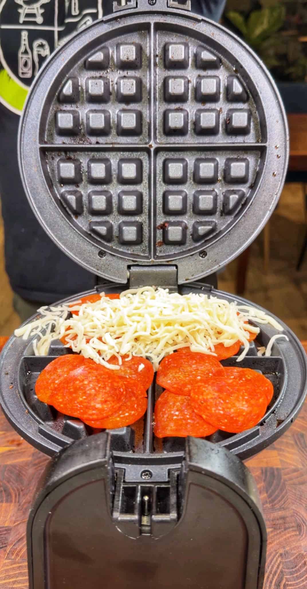 A waffle maker open to reveal slices of pepperoni and shredded cheese placed inside, ready to be cooked.