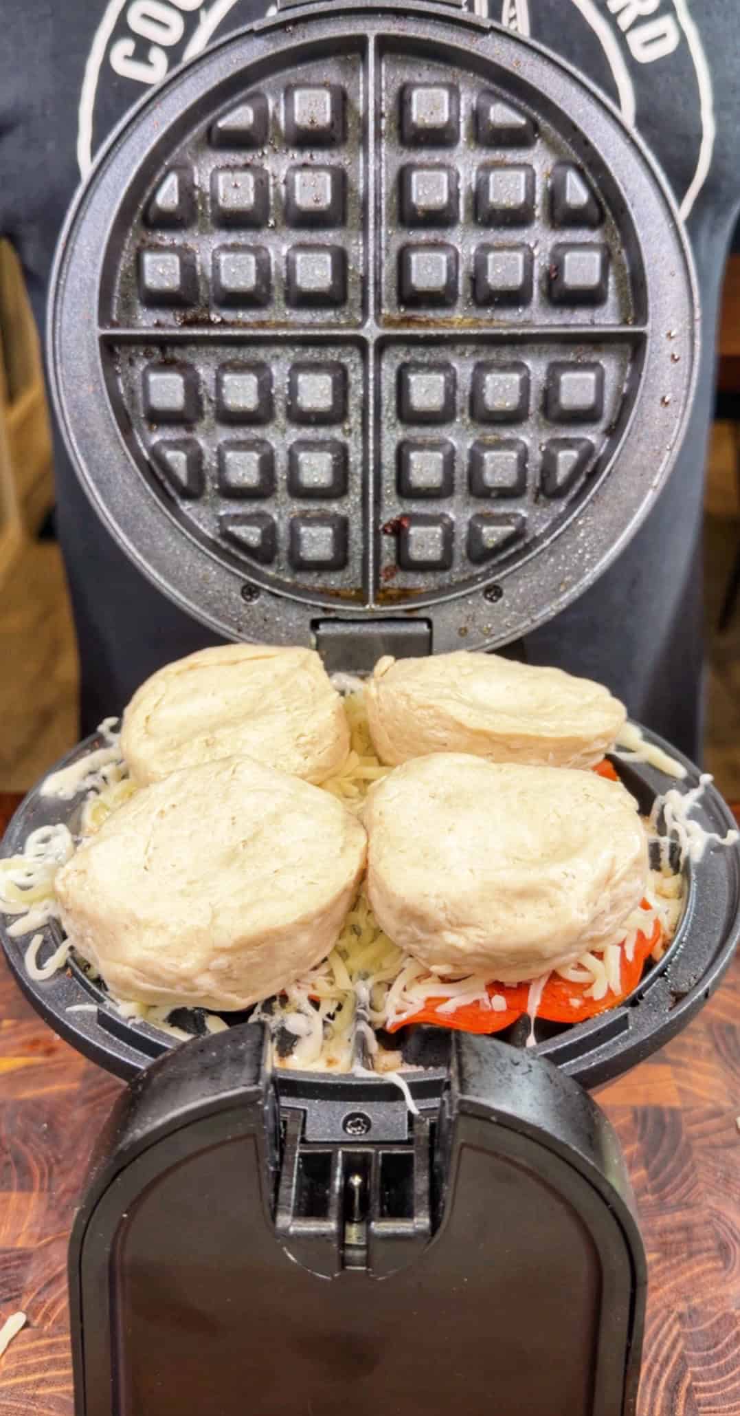 A close-up of an open waffle maker filled with shredded cheese, tomato slices, and four uncooked biscuit dough rounds, ready to be cooked.