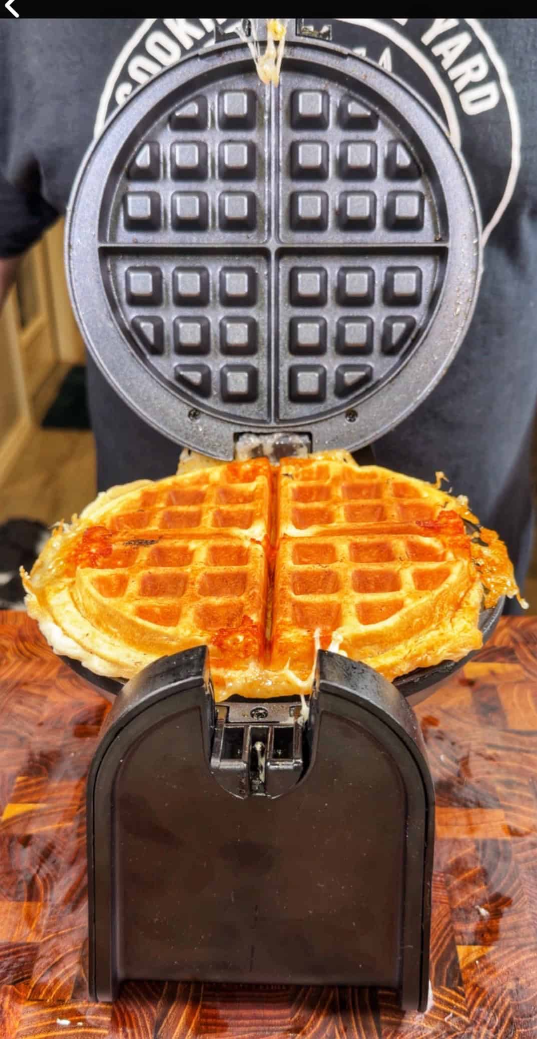 A golden, freshly cooked waffle sits in an open waffle maker on a wooden surface, with some melted cheese oozing out the sides. A person in a dark shirt stands in the background.