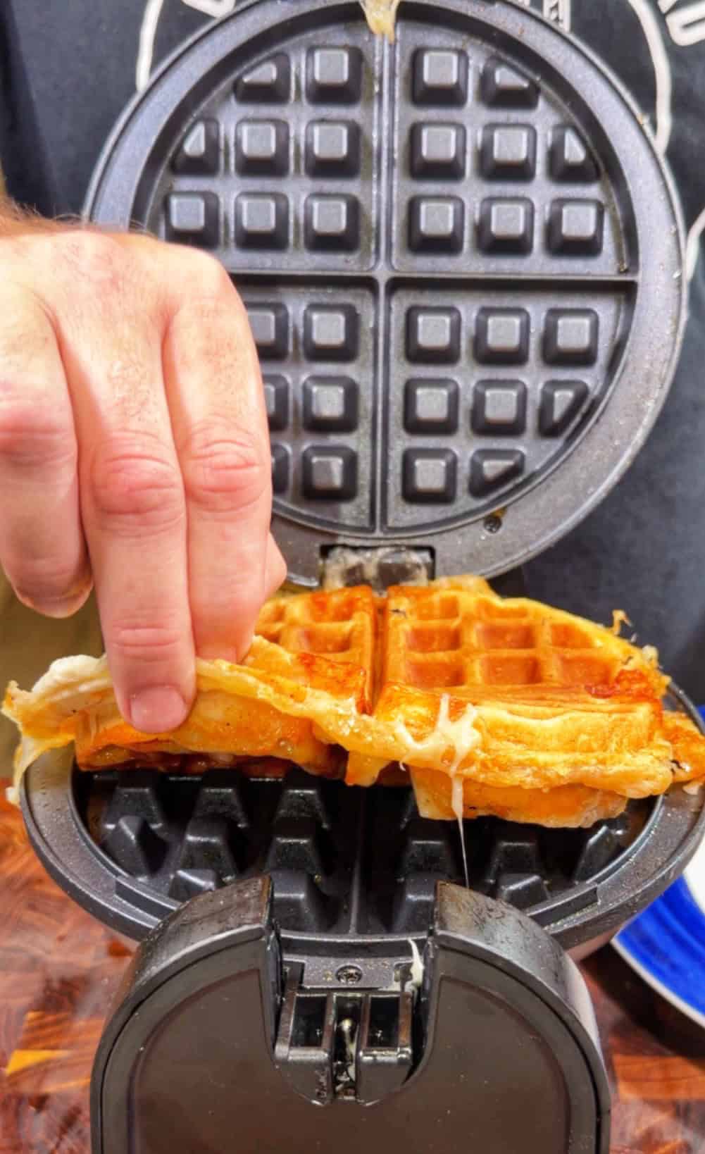 A hand lifts a cheesy, golden-brown waffle from an open waffle maker, with melted cheese stretching between the waffle and the appliance.