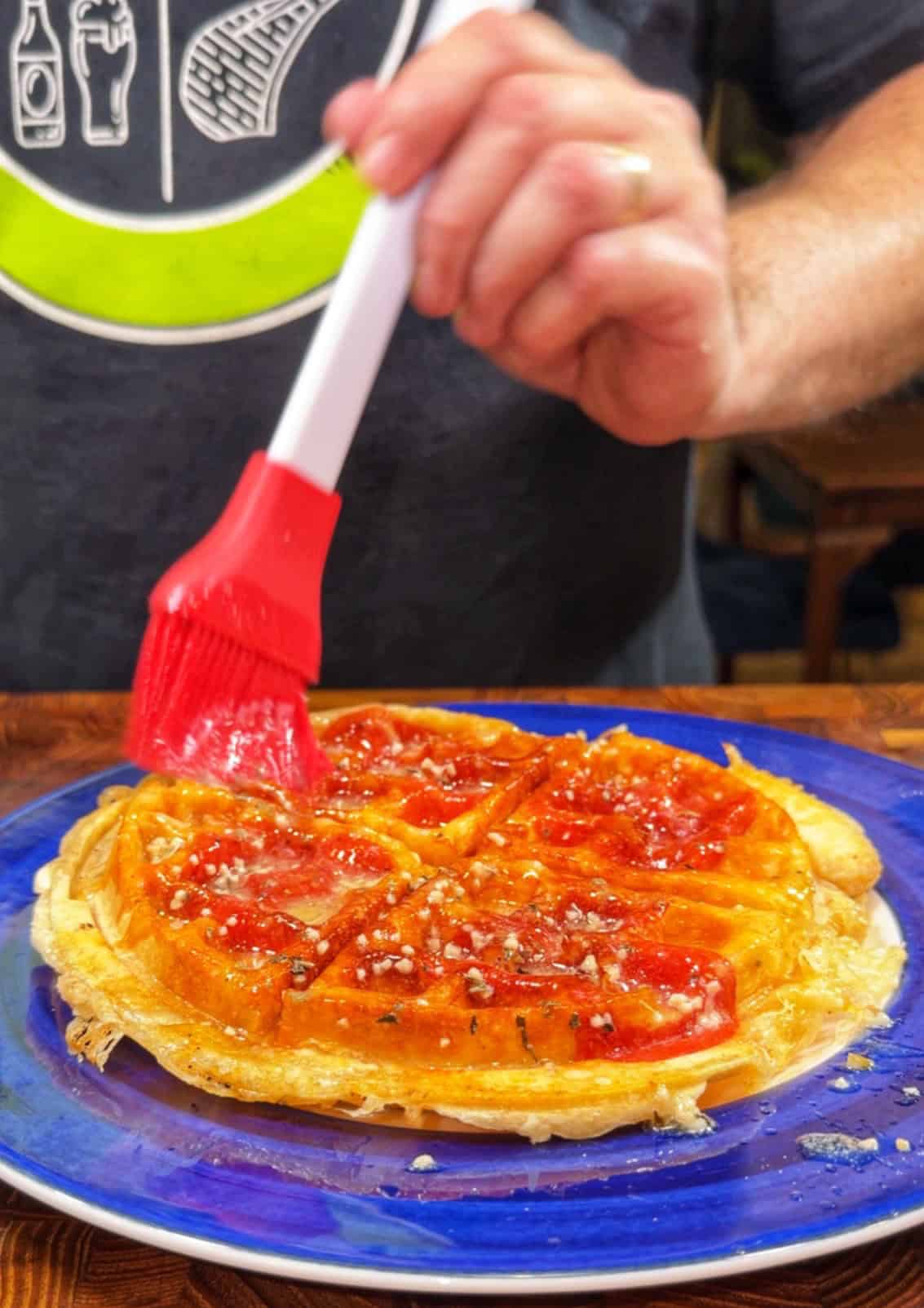 A person brushes sauce onto a golden, sliced waffle or pastry on a blue plate, using a red silicone brush. The pastry is topped with sauce and sprinkled seasoning. The person wears a dark shirt with a graphic design.