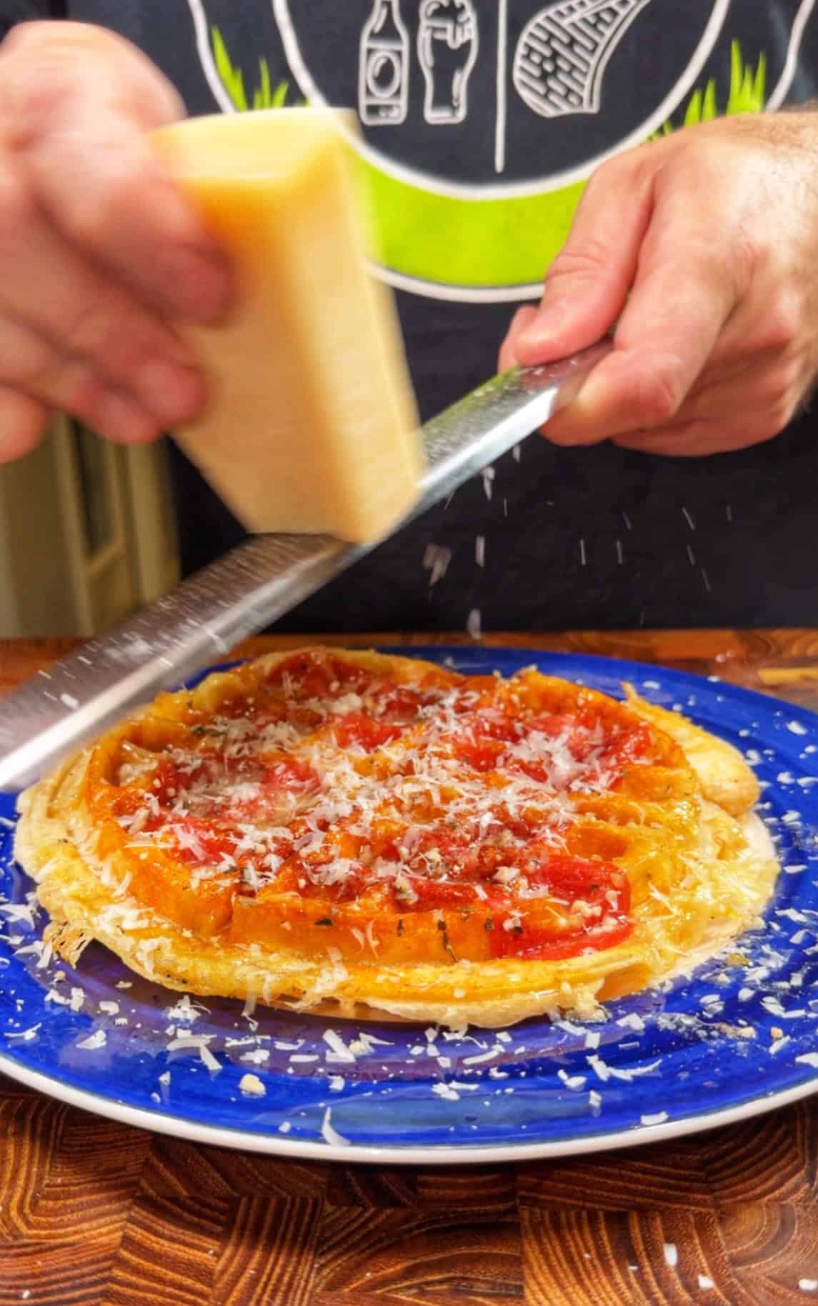 A person grates cheese over a tomato-covered waffle on a blue plate, with cheese shavings falling onto the waffle. The persons torso is visible, wearing a dark shirt with white graphics.