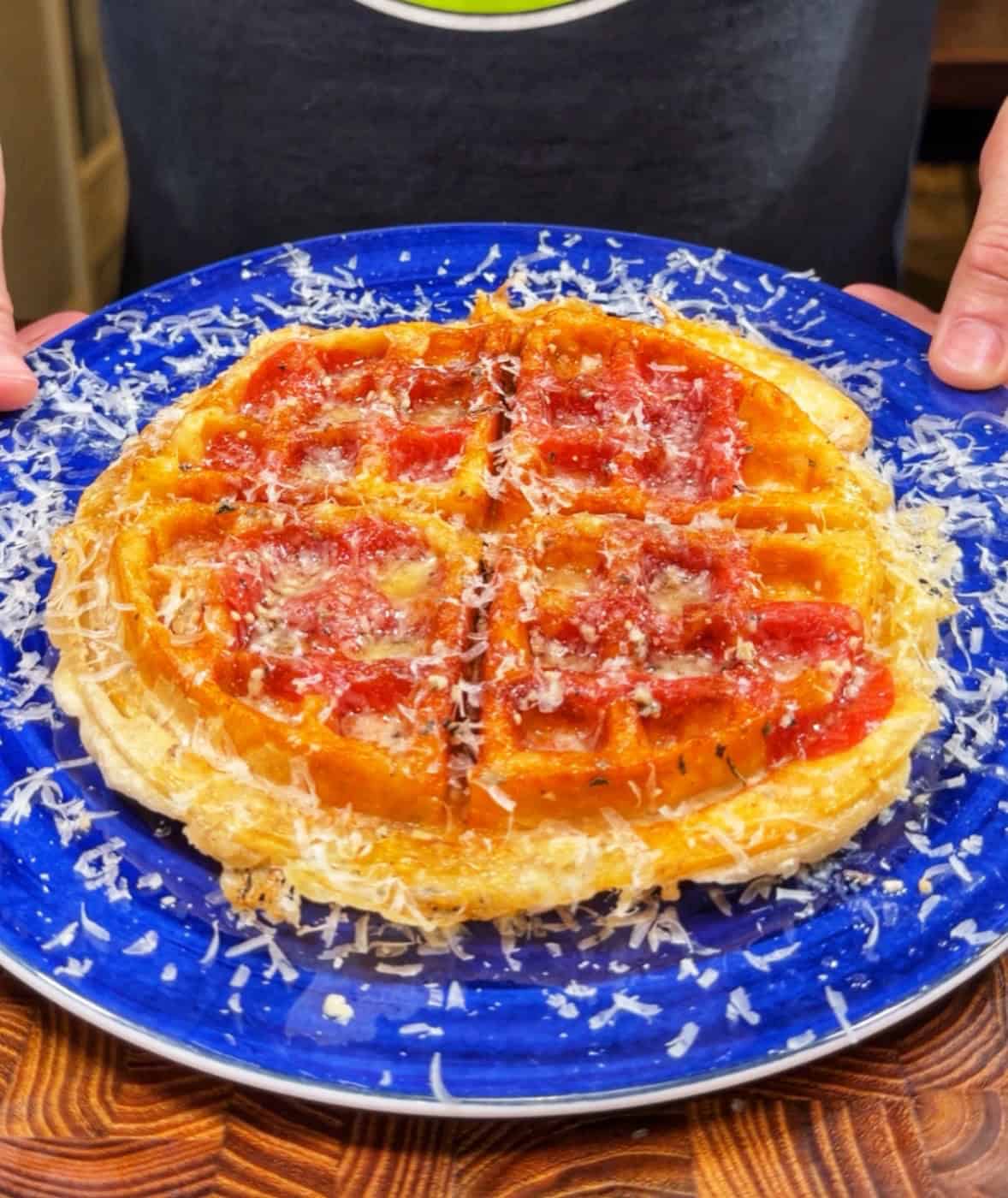 A person holds a blue plate with a golden waffle topped with tomato sauce and shredded cheese, resembling a pizza. The waffle is divided into four sections and served on a wooden surface.