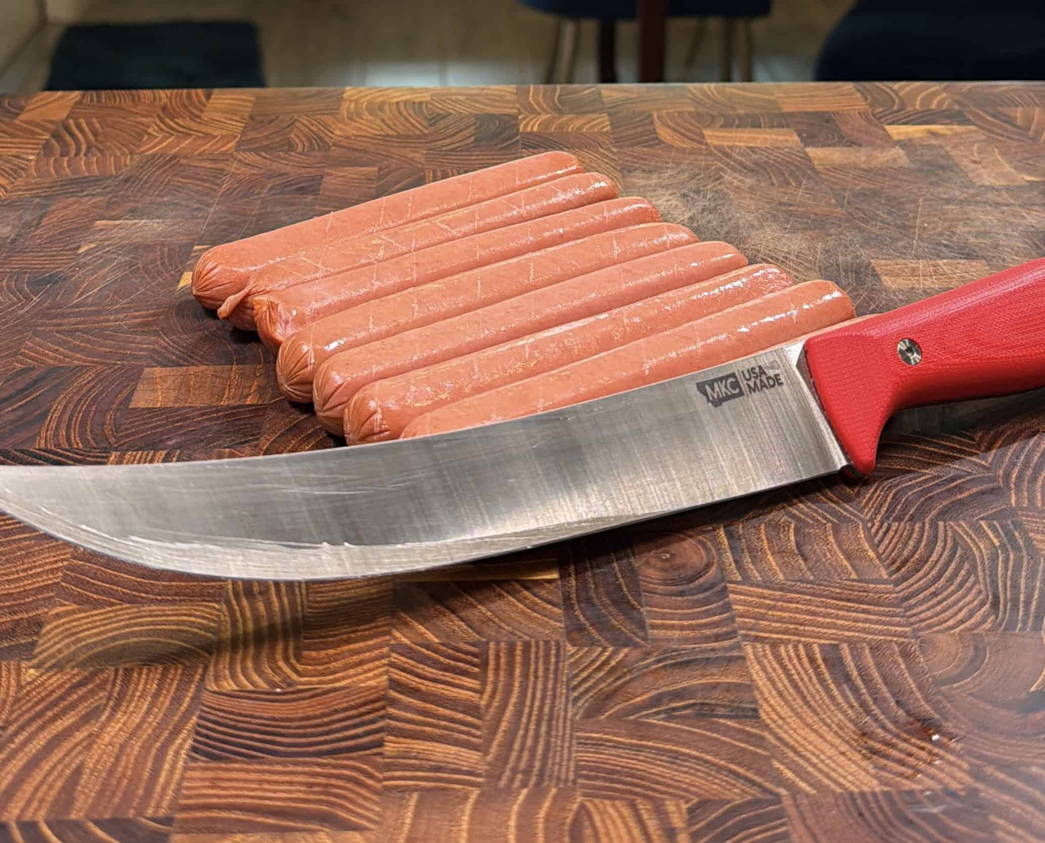 A large chefs knife with a red handle rests on a wooden cutting board next to a stack of eight uncooked hot dogs still in their casings.