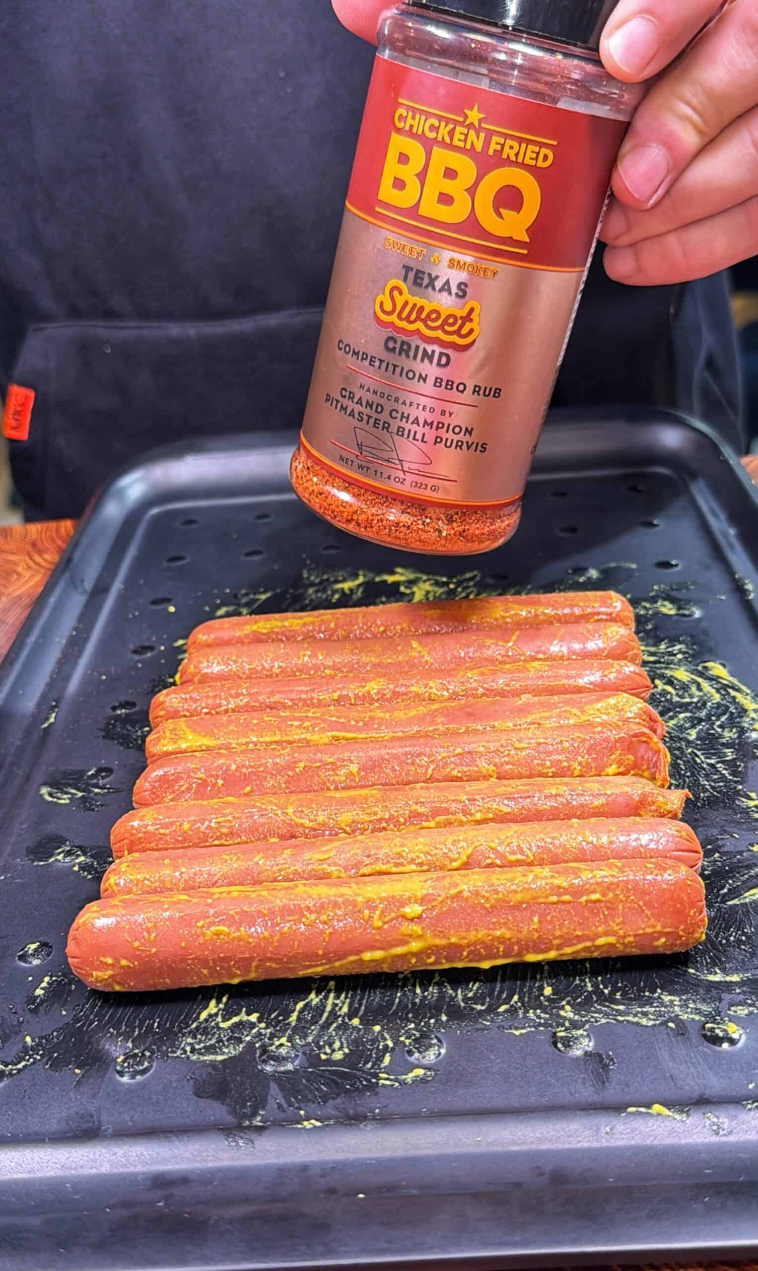 A person sprinkles Texas Sweet BBQ rub onto a tray of uncooked hot dogs, which are coated in mustard and arranged neatly on a black baking sheet.