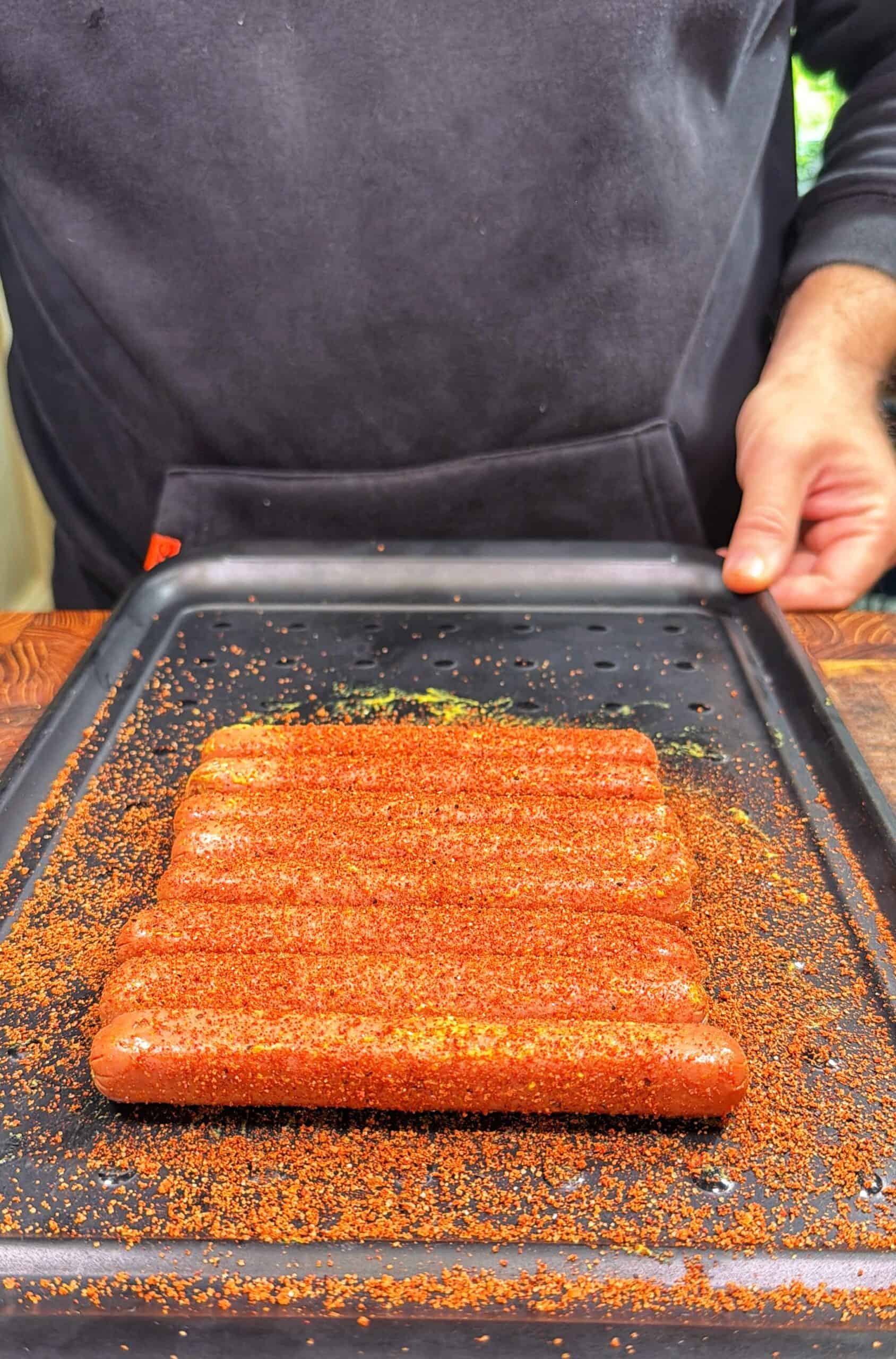 A person in a dark sweatshirt holds a tray of six hot dogs coated with a reddish seasoning, ready for grilling or cooking.