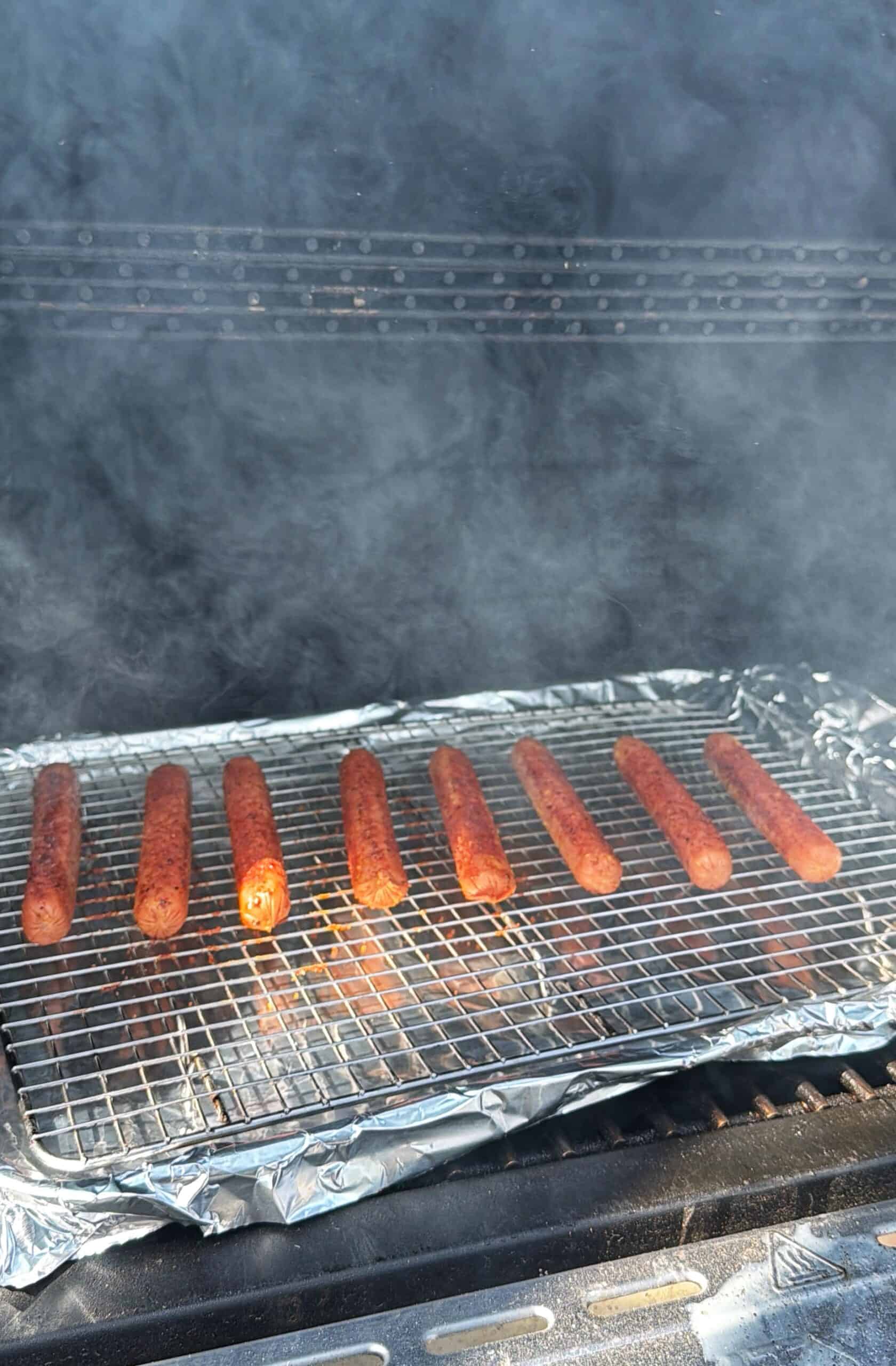 Seven hot dogs are lined up on a wire rack covered with foil inside a smoky grill. Smoke wafts around, partially obscuring the background.