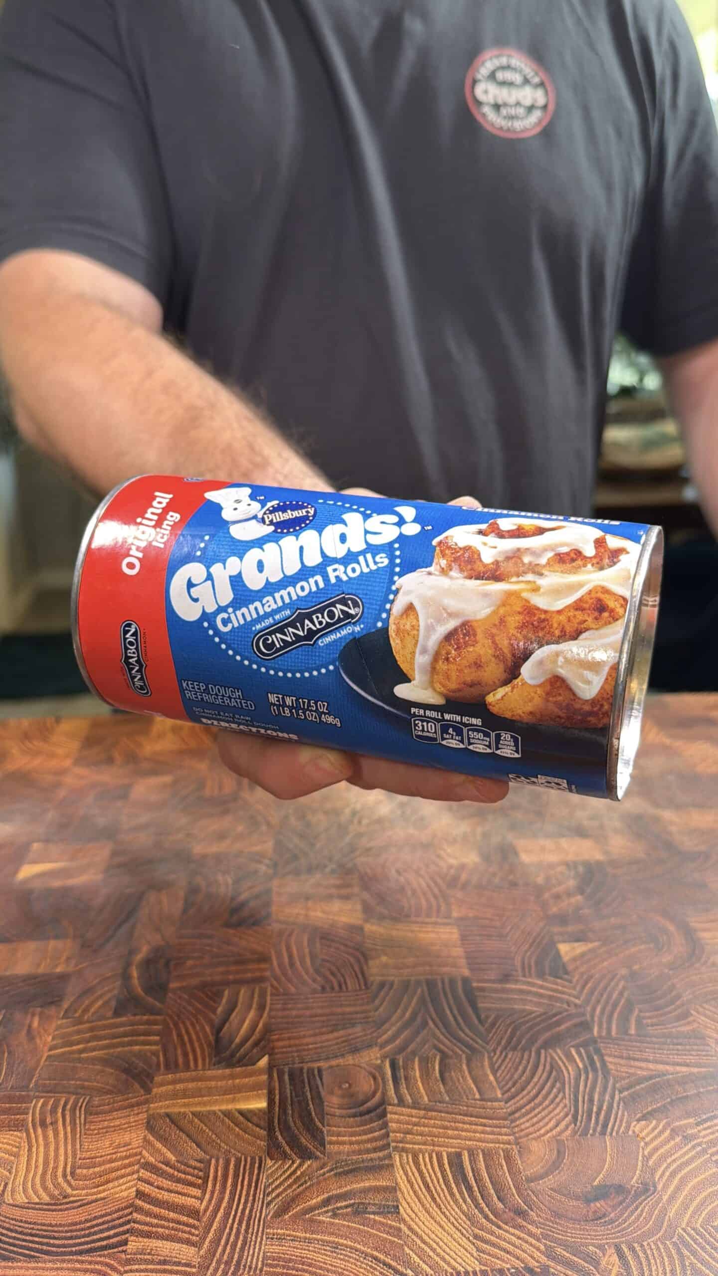 A person holds a can of Pillsbury Grands! Cinnamon Rolls with icing above a wooden countertop. Only the persons arm and part of their torso are visible, wearing a dark shirt with a logo.