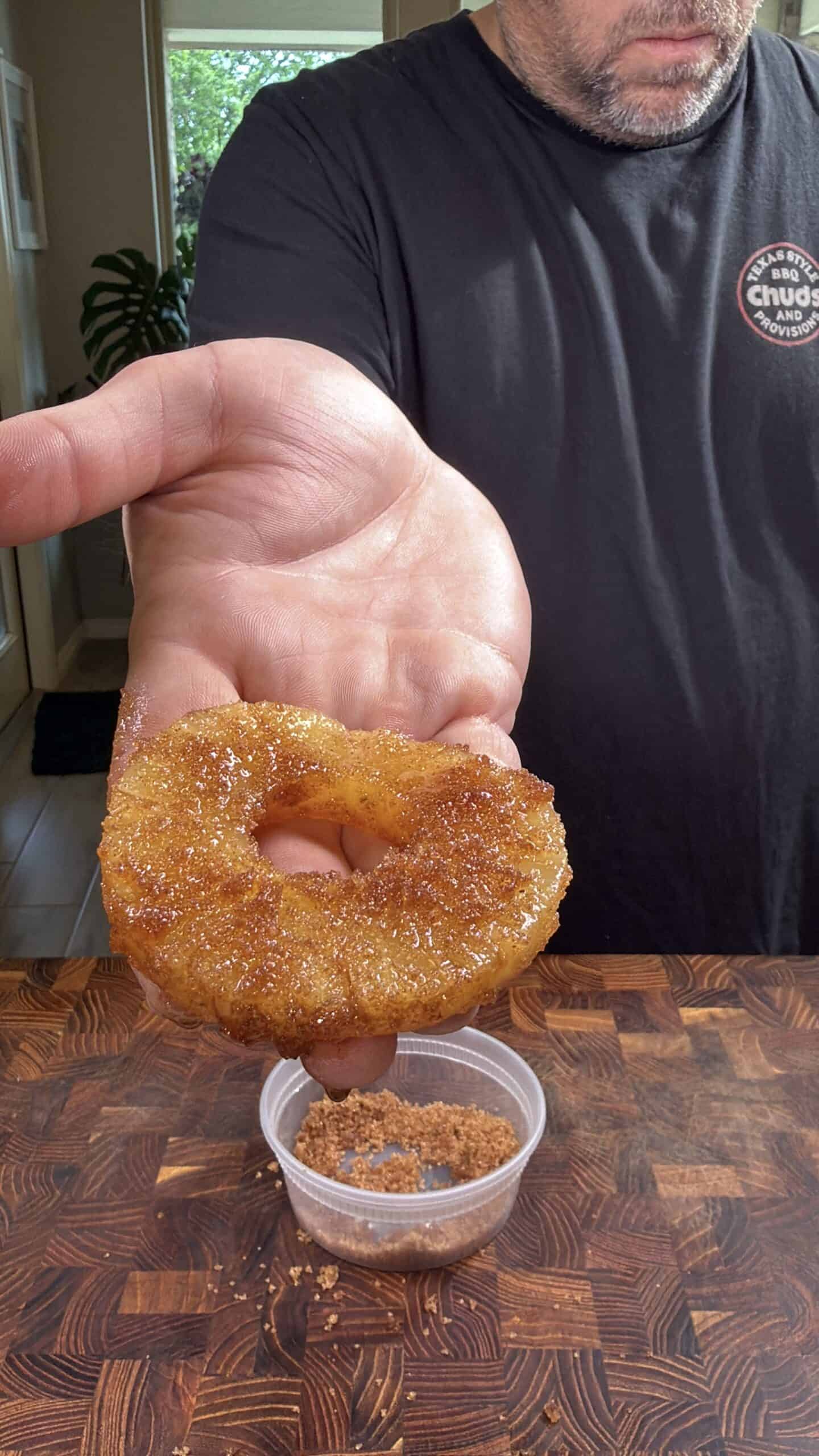 A person holds a caramelized pineapple ring in their hand over a wooden surface, with a small container of brown sugar or seasoning below. The person is wearing a black T-shirt.