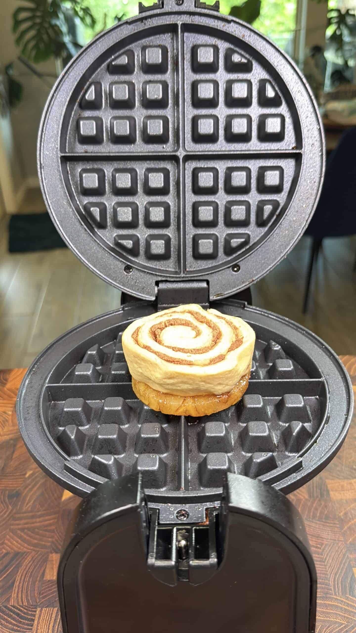 A single cinnamon roll sits in the center of an open waffle maker, ready to be pressed. The waffle maker is on a wooden surface with plants visible in the background.