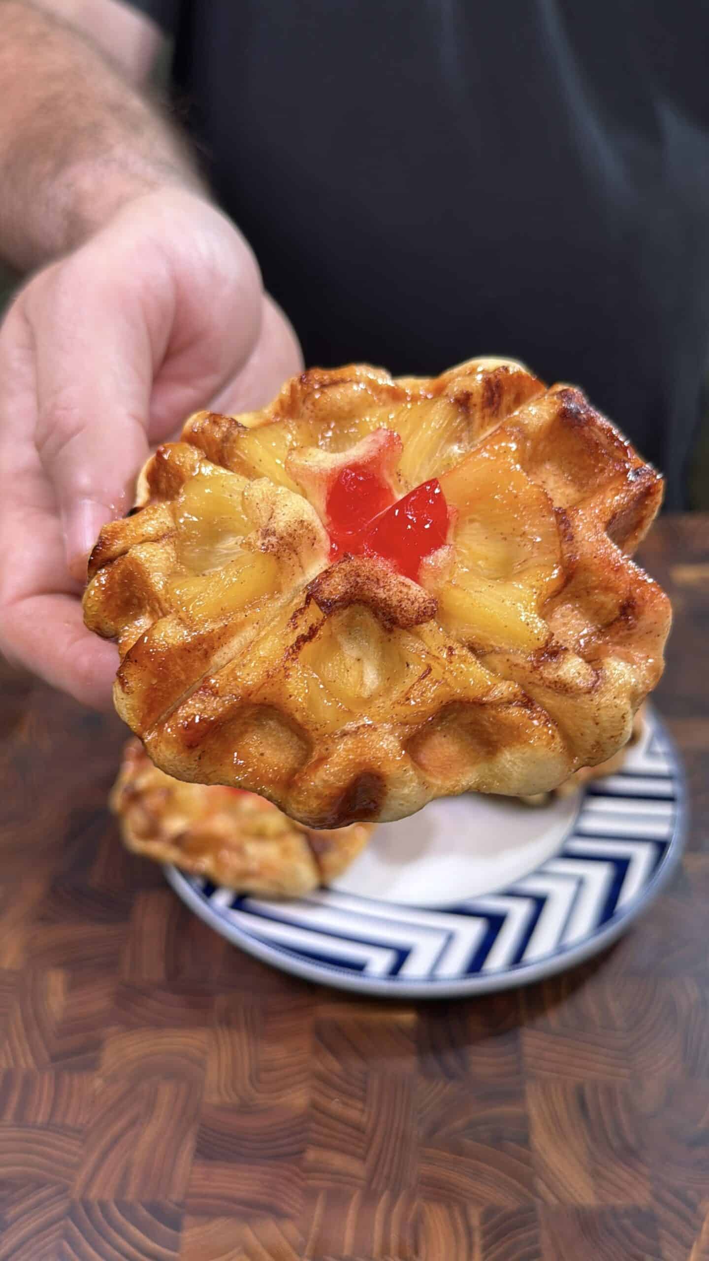 A hand holding a pastry topped with pineapple slices and a red cherry, with another pastry on a blue and white plate in the background. The pastries are on a wooden surface.