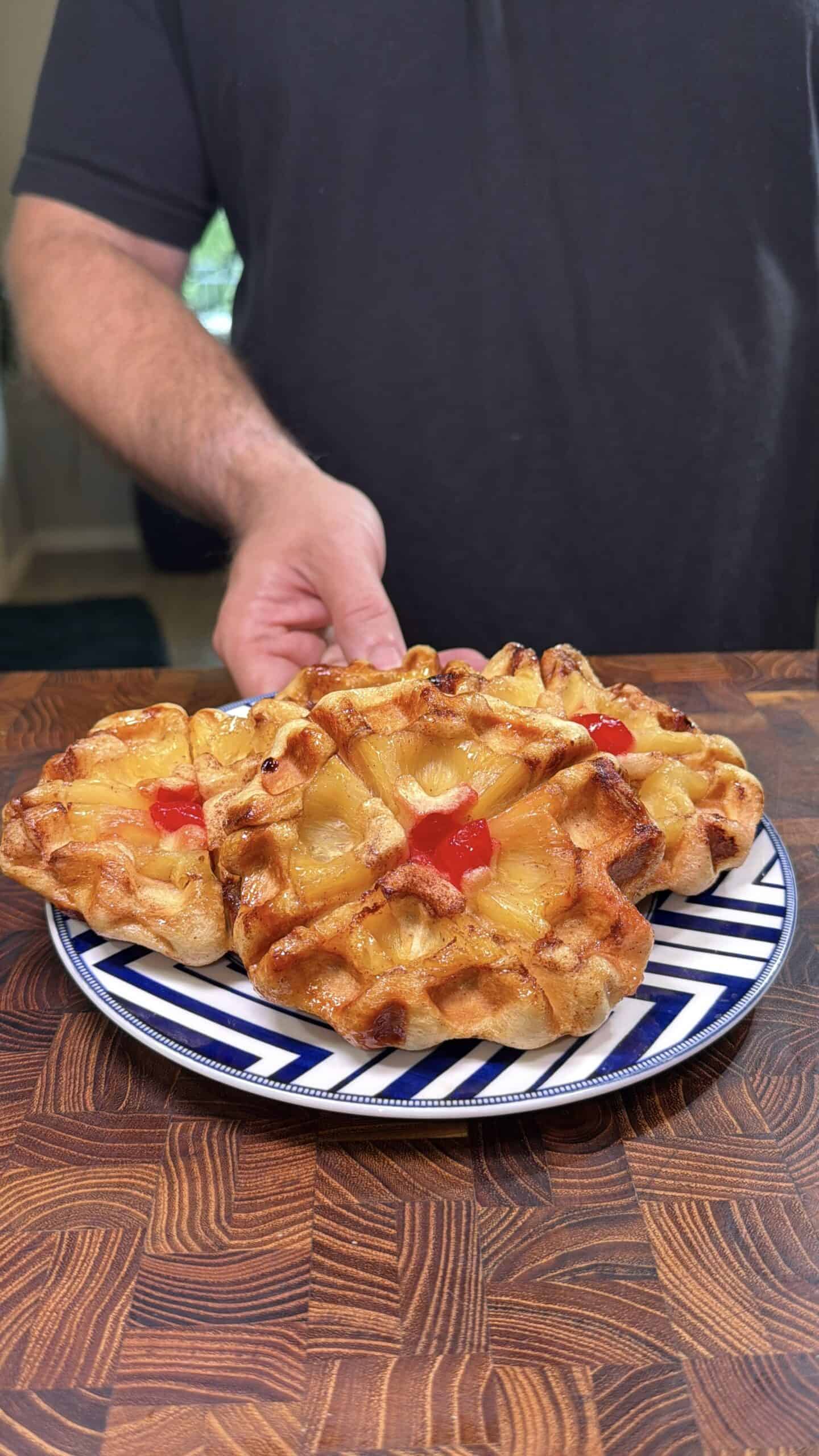 A person holding a plate with three pineapple upside-down waffles topped with pineapple rings and cherries, on a wooden surface.