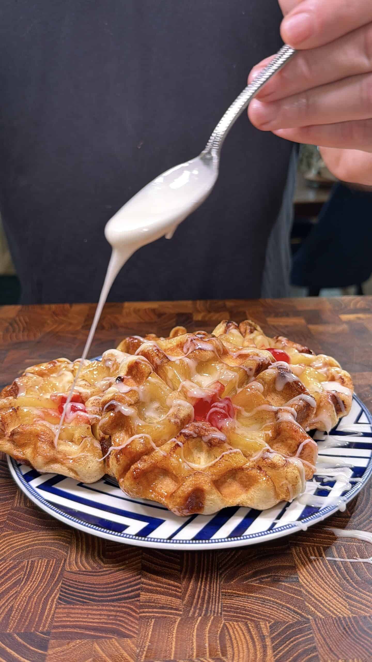A hand holds a spoon drizzling white icing over a plate of golden brown waffles topped with fruit, on a patterned plate set on a wooden table.