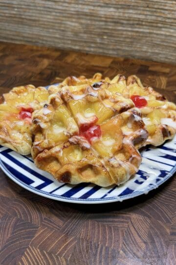 Three pineapple danish pastries with cherries and icing are arranged on a blue and white plate, sitting on a wooden surface with a textured background.