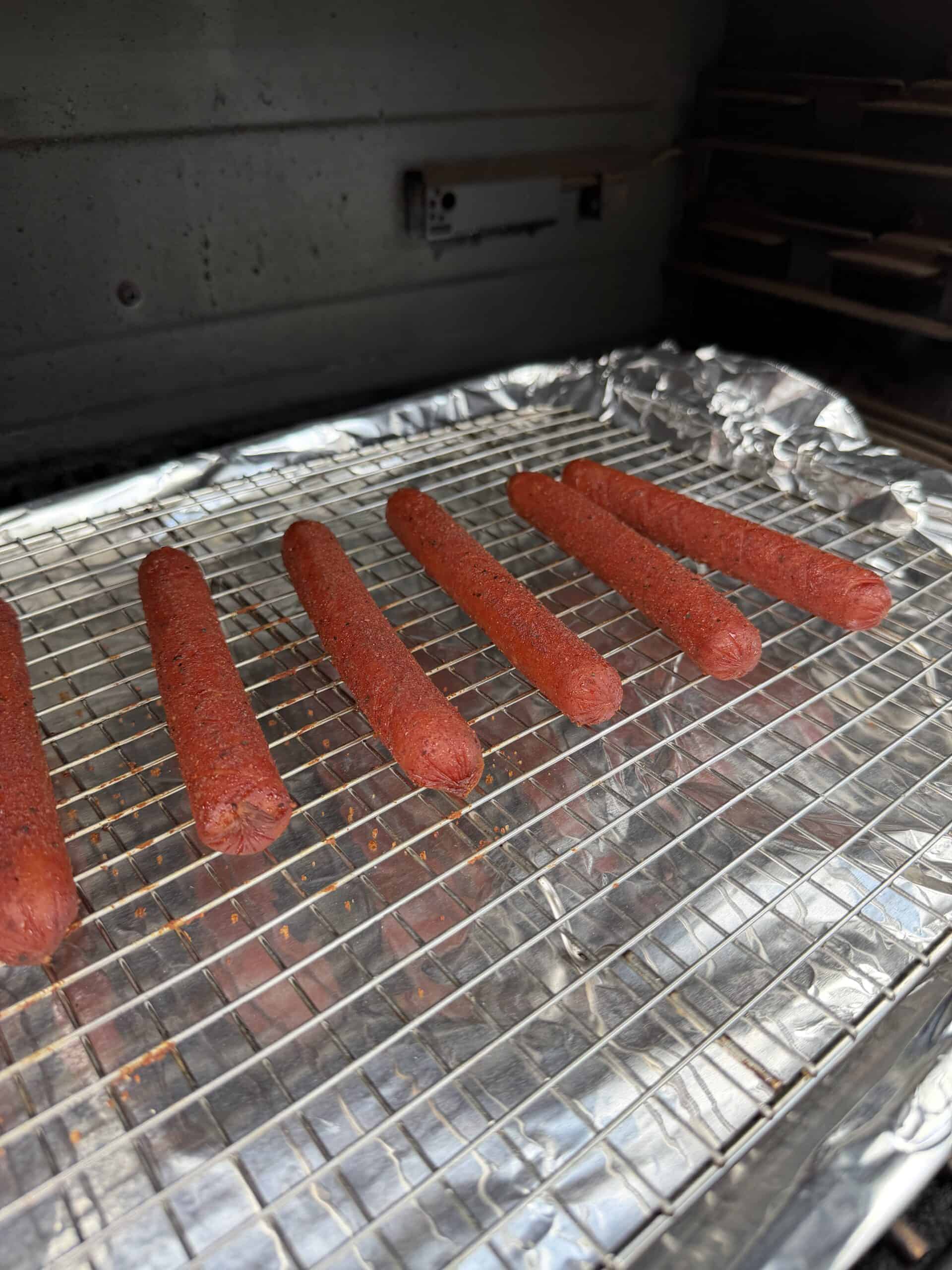 Five seasoned hot dogs are arranged on a wire rack over a foil-lined baking tray, inside an oven or smoker.