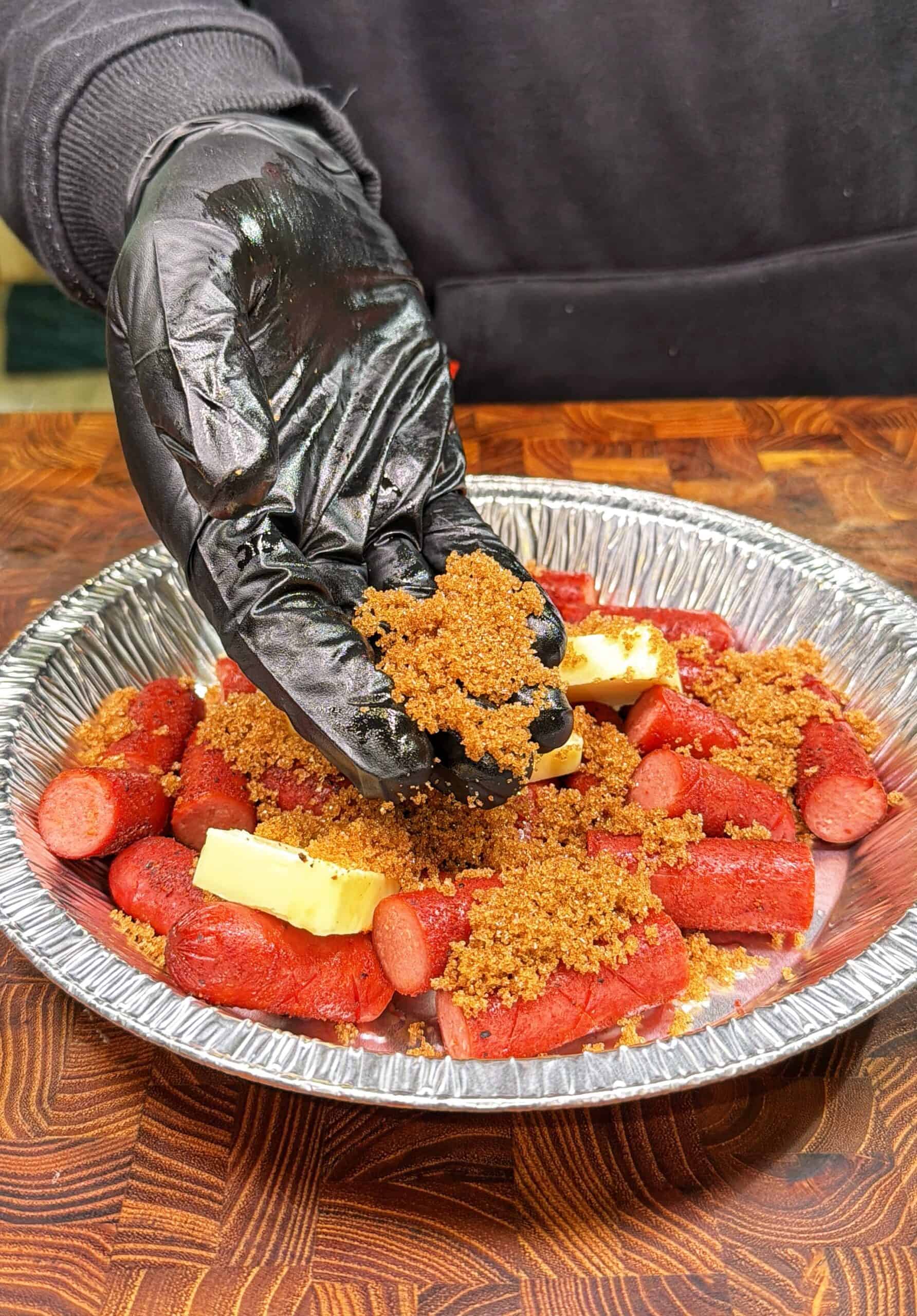 A gloved hand sprinkles brown sugar over sliced hot dogs and butter pieces in a disposable aluminum pie pan on a wooden surface.