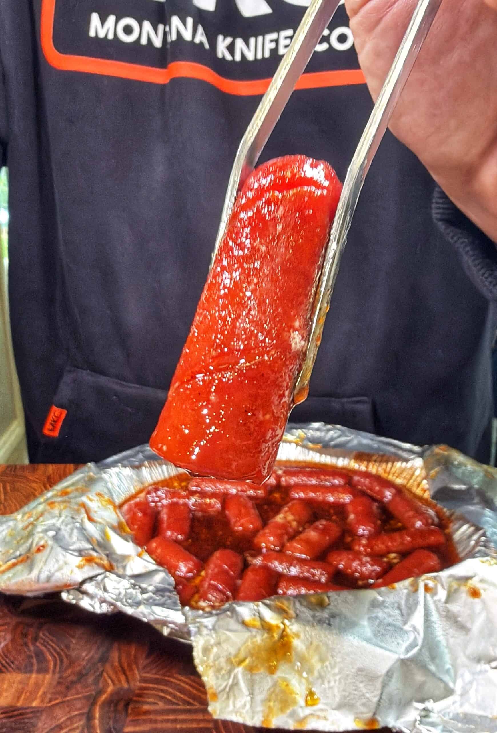 A person uses metal tongs to hold a sauce-covered sausage over a foil tray filled with more sausages in sauce, while wearing a dark sweatshirt with a Montana Knife logo.