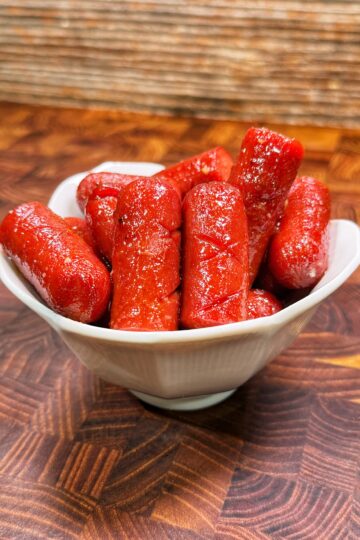 A white bowl filled with bright red, shiny cocktail sausages sits on a patterned wooden surface, with a textured brown background behind it.