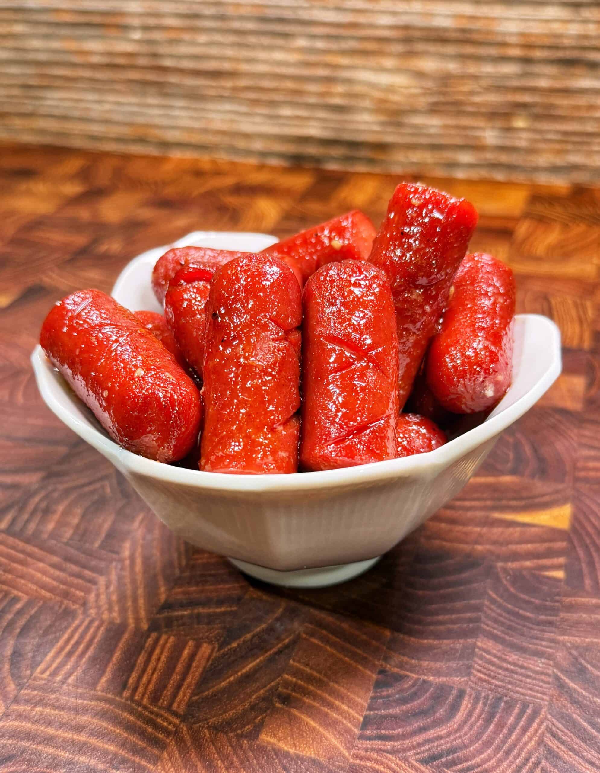 A white bowl filled with bright red, shiny cocktail sausages sits on a patterned wooden surface, with a textured brown background behind it.