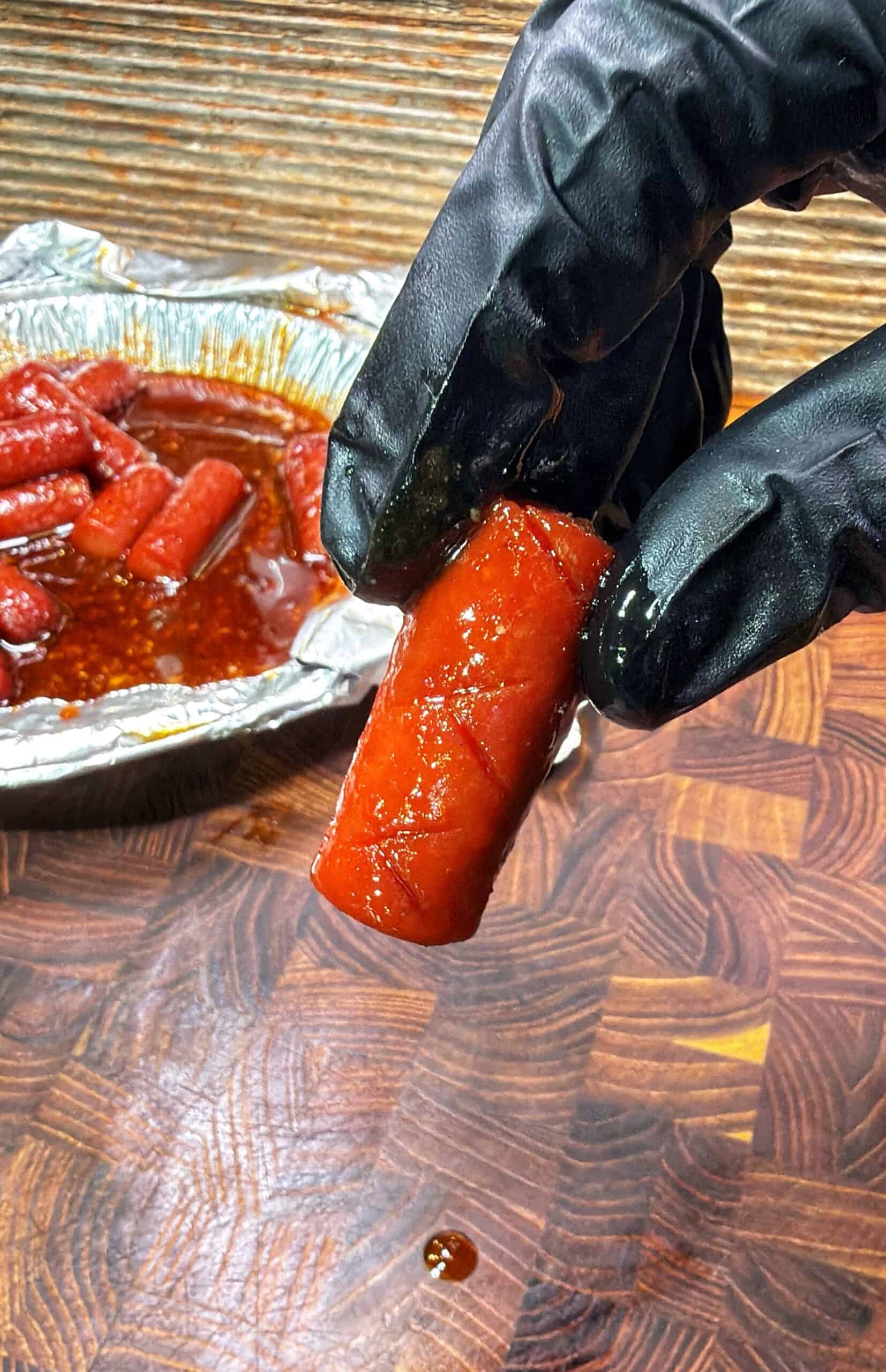 A gloved hand holds a shiny, sauce-covered smoked sausage above a wooden surface, with a foil tray of more sausages in sauce in the background.
