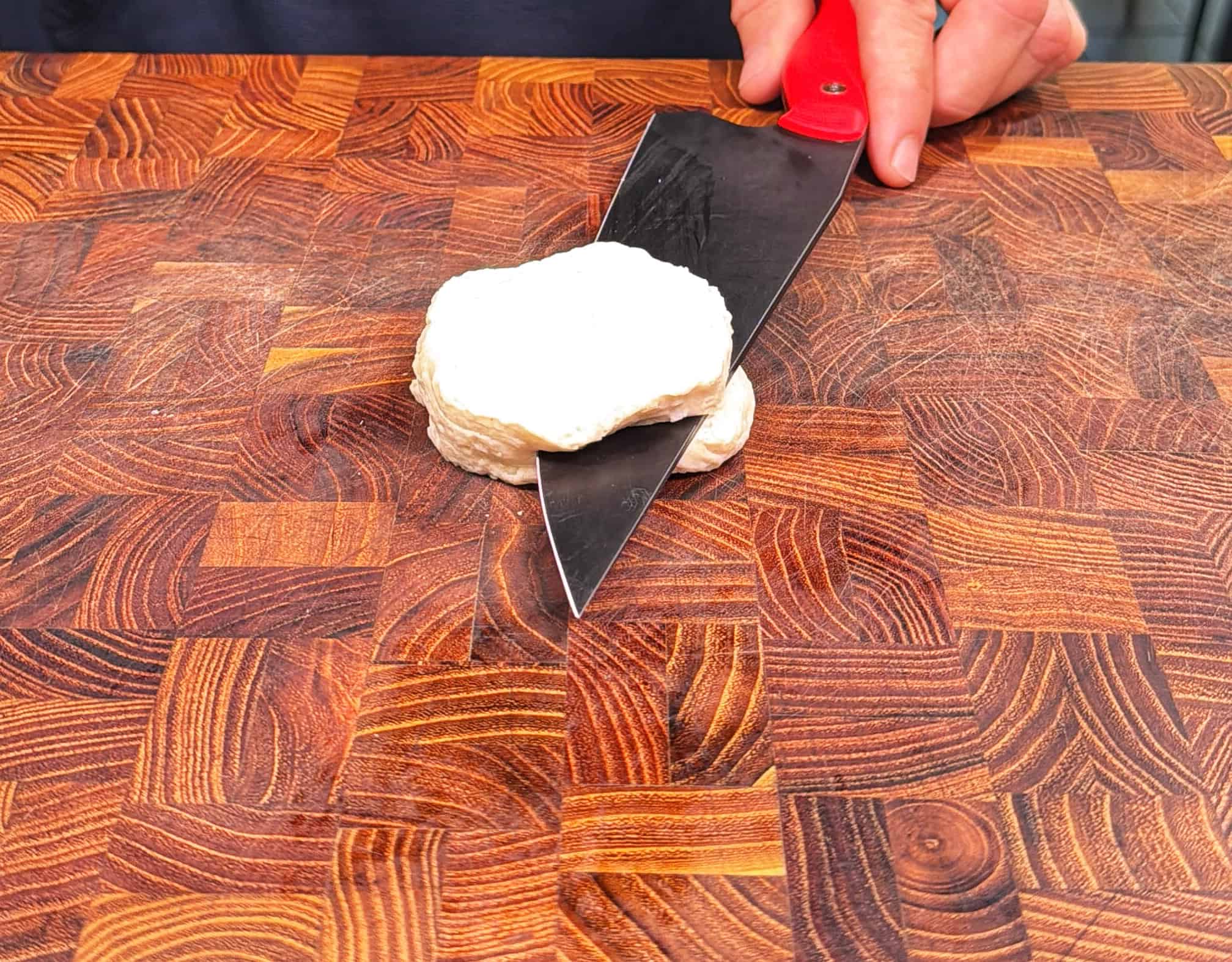 A hand holding a knife slices through a round piece of uncooked biscuit dough on a patterned wooden cutting board.