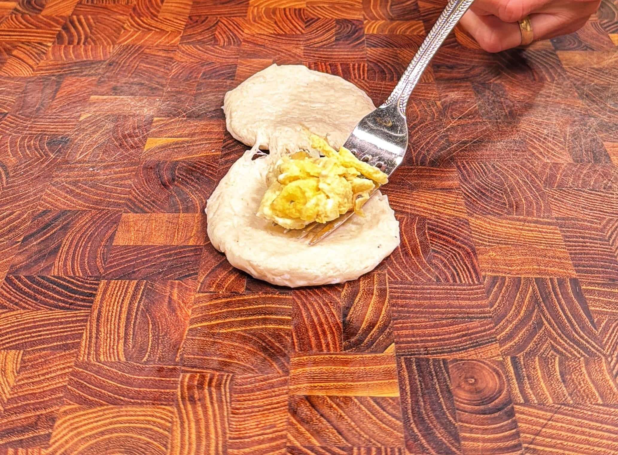 A hand uses a fork to place a scrambled egg filling onto a round piece of dough on a wooden patterned surface, preparing to fold it closed.