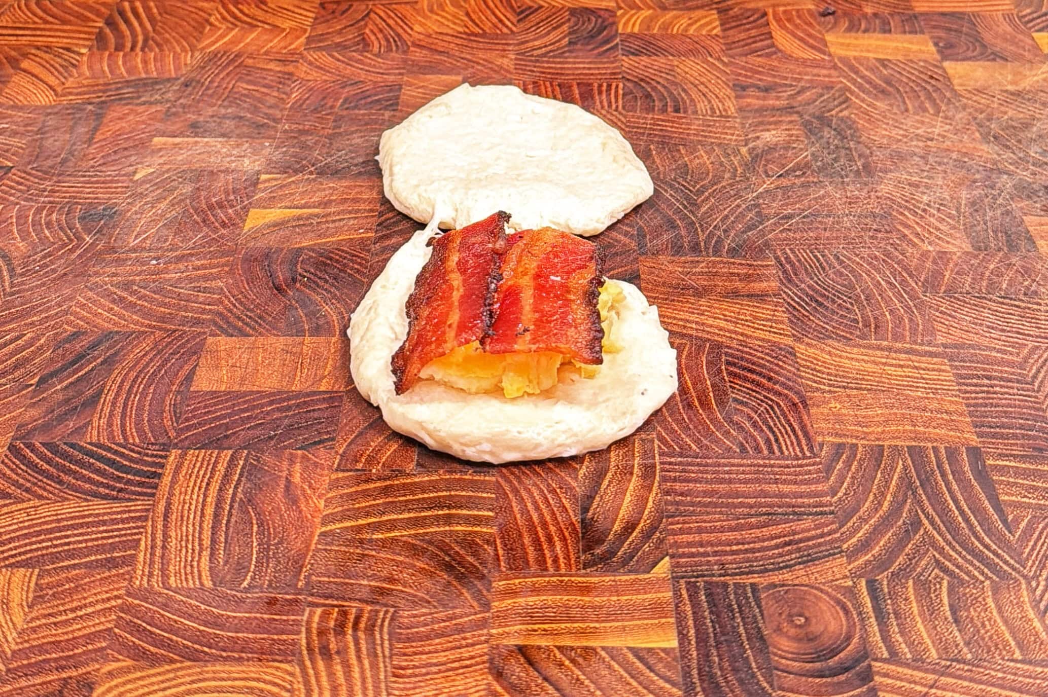 A piece of uncooked dough topped with slices of cooked bacon and cheese sits on a wooden cutting board, with another piece of plain dough above it.
