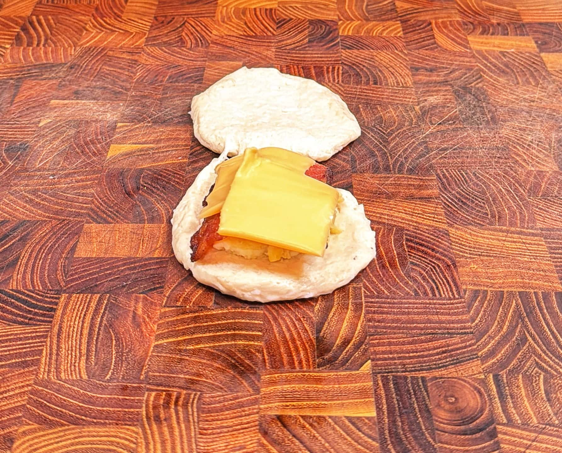 An open biscuit on a wooden surface, topped with slices of cheddar cheese and what appears to be bacon or ham. The biscuit top is placed behind the bottom half.