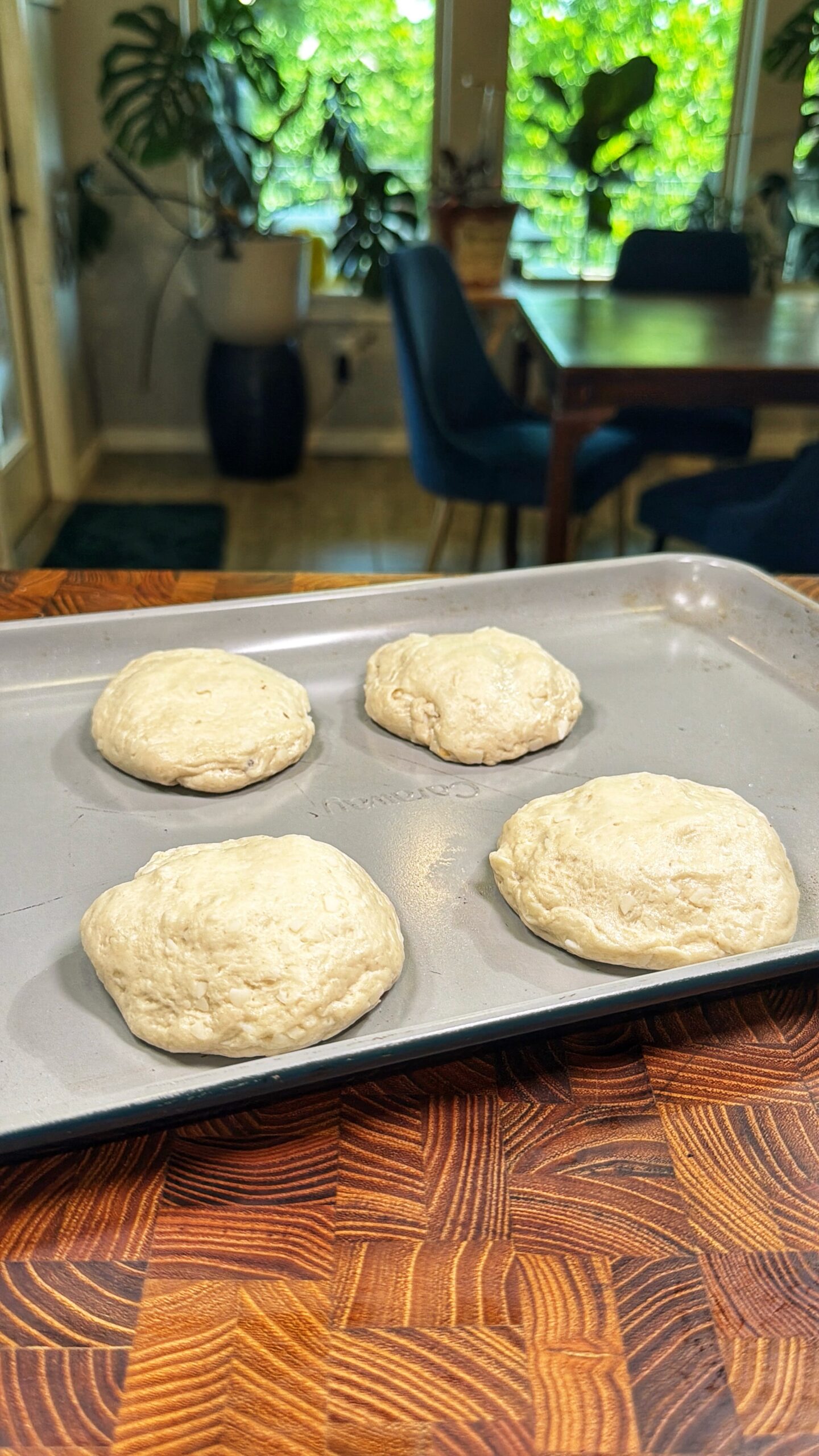 Four unbaked pizza dough rounds are placed on a baking sheet, sitting on a wooden countertop. In the background, there is a dining table, blue chairs, houseplants, and large windows with greenery outside.
