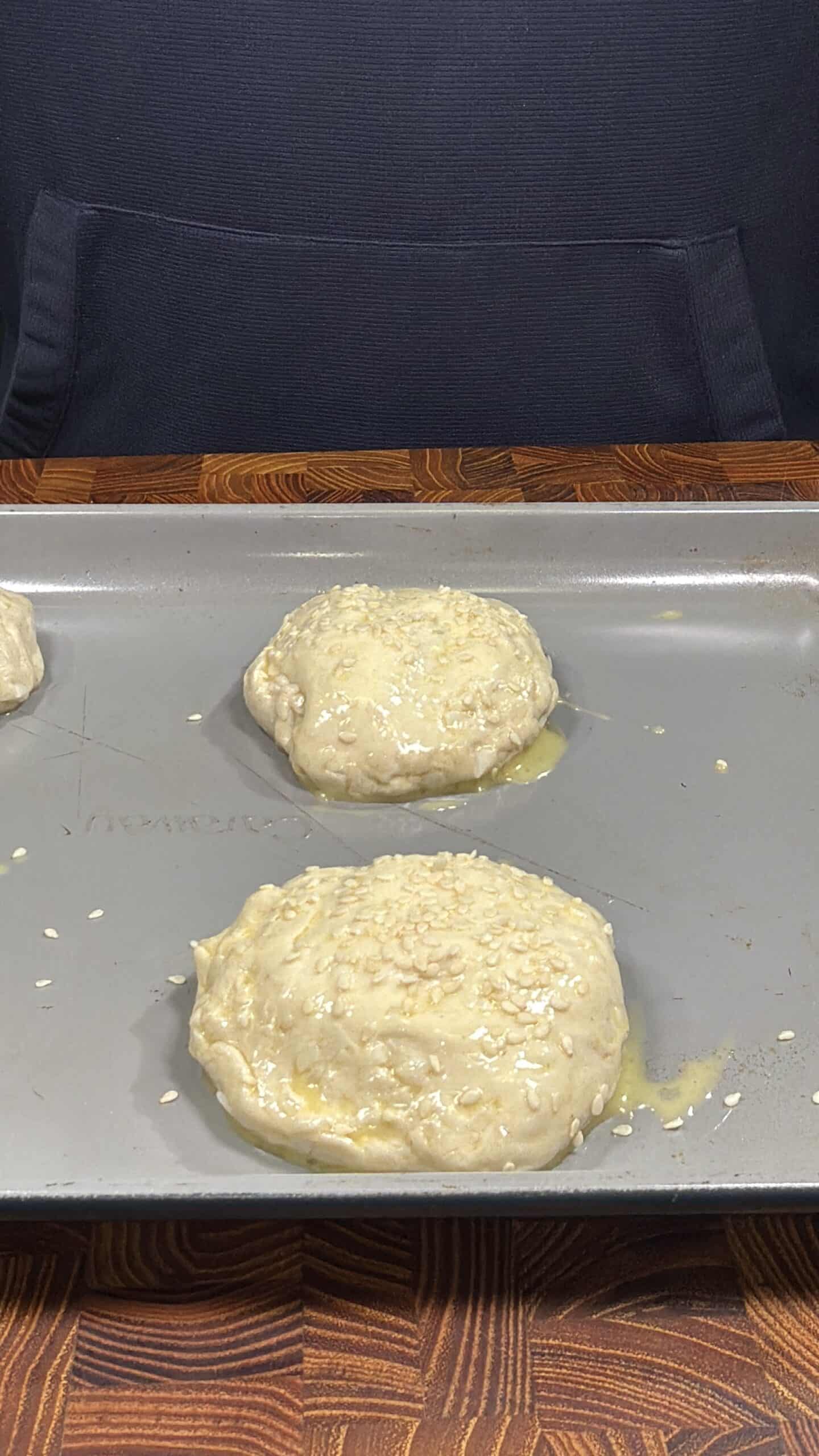 Unbaked, round dough patties topped with sesame seeds and brushed with egg wash on a baking sheet, ready to be baked. A dark background and wooden surface are partially visible.