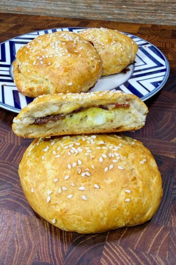 A close-up of golden, sesame-topped baked buns. One bun is sliced open to reveal a cheesy and savory filling, while more whole buns sit on a decorative plate in the background.