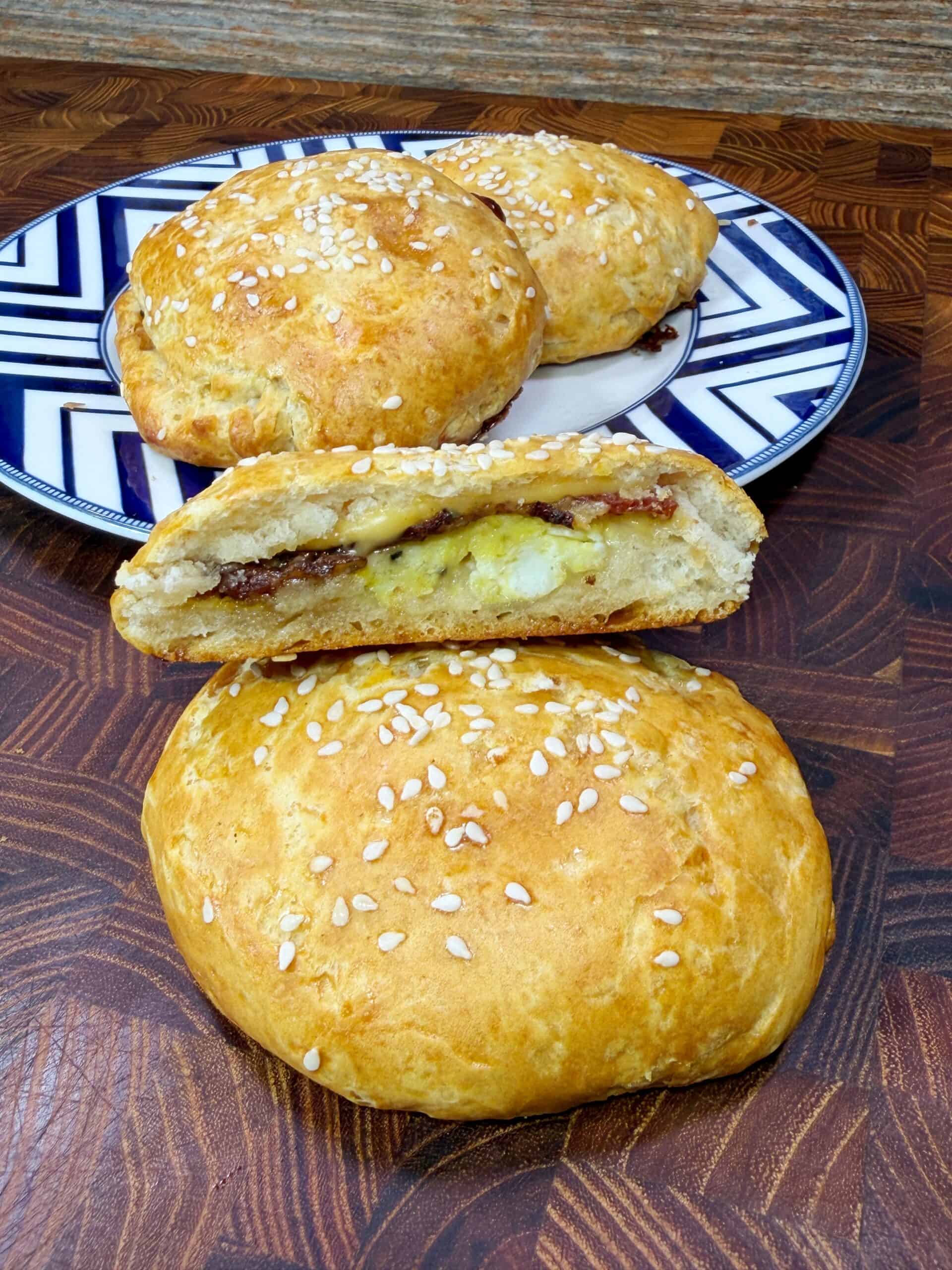 A close-up of golden, sesame-topped baked buns. One bun is sliced open to reveal a cheesy and savory filling, while more whole buns sit on a decorative plate in the background.