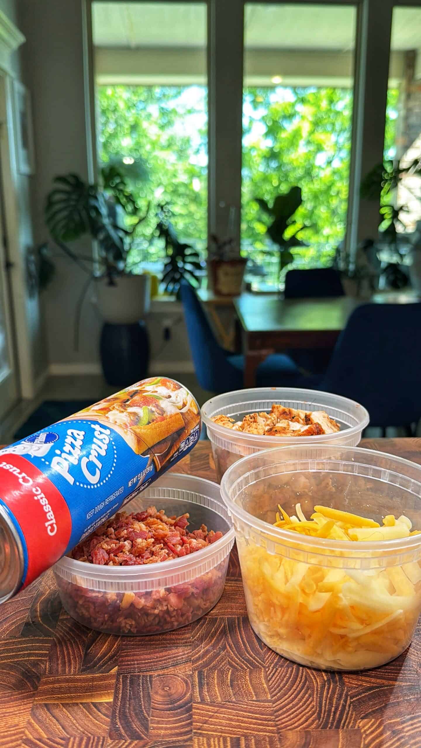 A can of Pillsbury pizza crust sits on a wooden table next to plastic containers filled with cooked bacon bits, shredded cheese, and cooked chicken, with a bright dining room and green plants in the background.