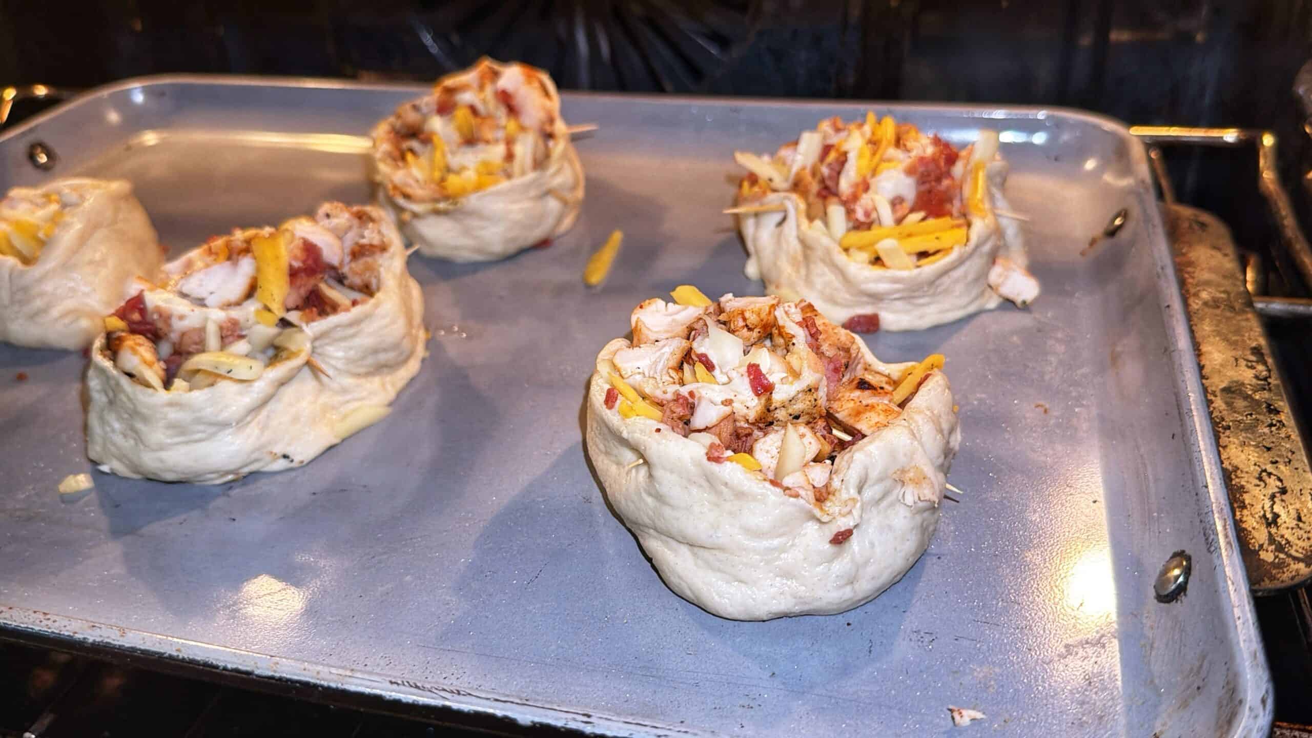 Five unbaked bread bowls filled with cheese, bacon, and seasoned ingredients are arranged on a metal baking sheet inside an oven, ready to be baked.