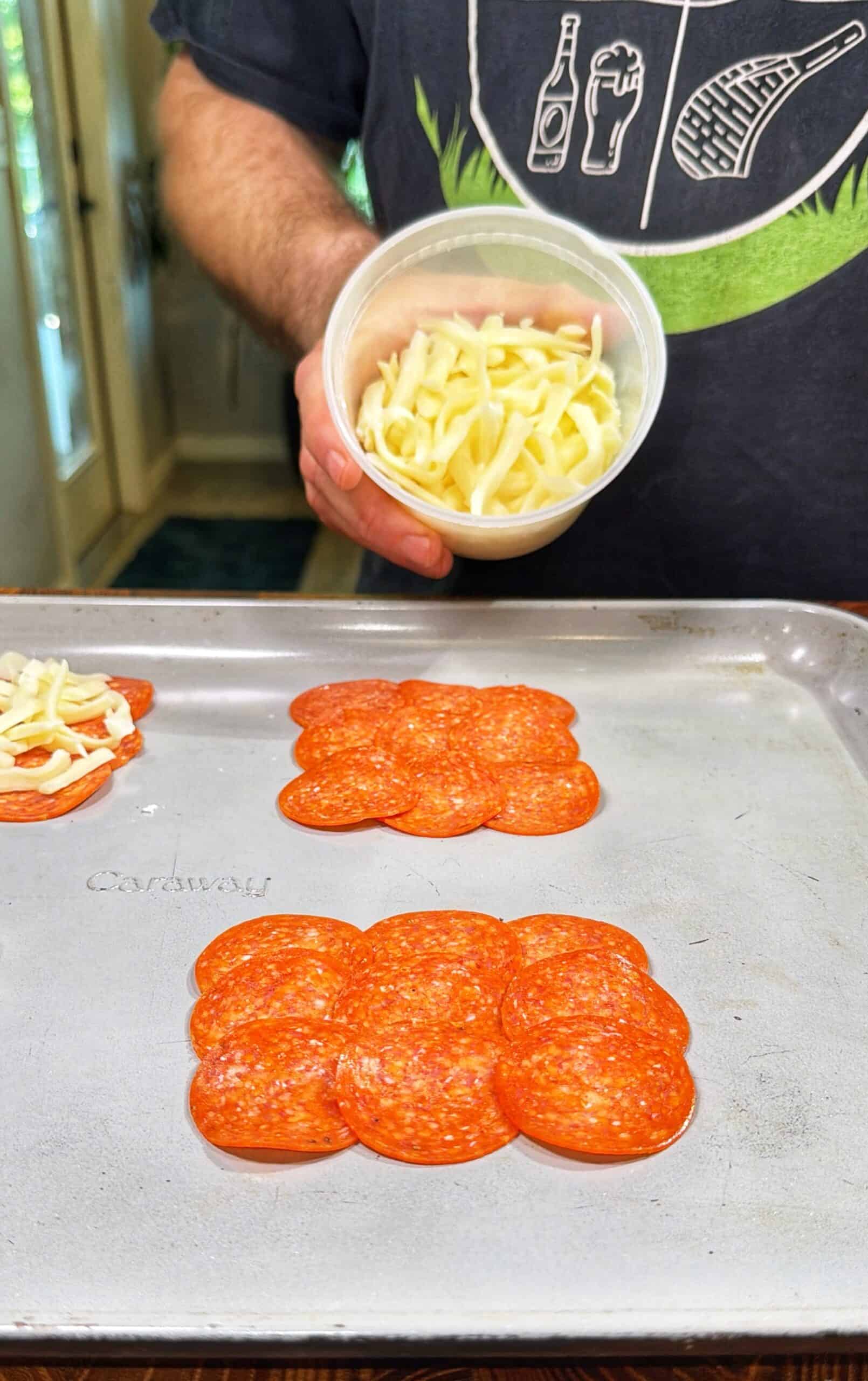 A person in a black shirt holds a container of shredded cheese above a baking tray with arranged slices of pepperoni, preparing to add cheese.