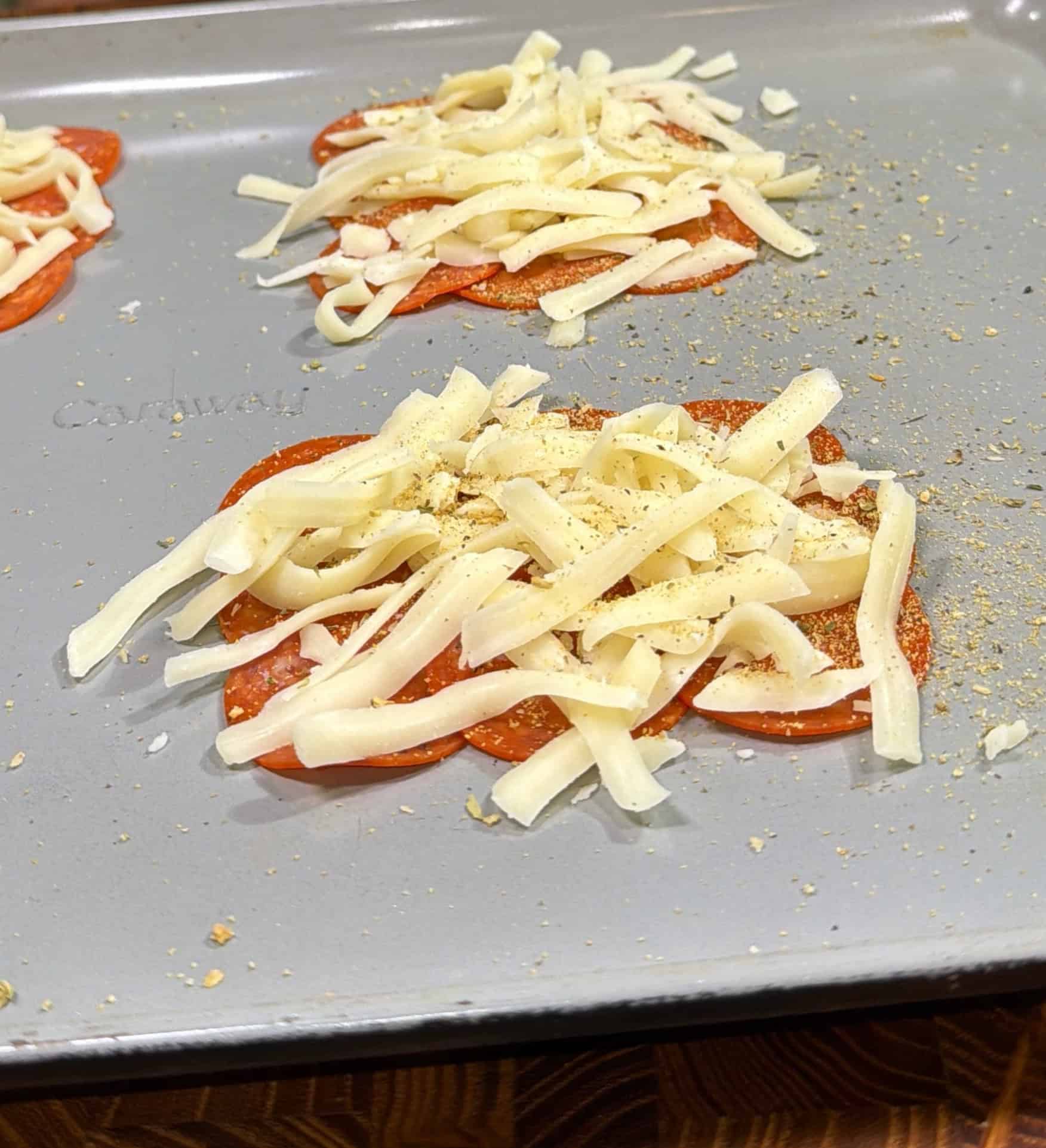 Slices of pepperoni arranged in circles on a baking sheet, topped with shredded cheese and sprinkled with seasoning, ready to be baked.