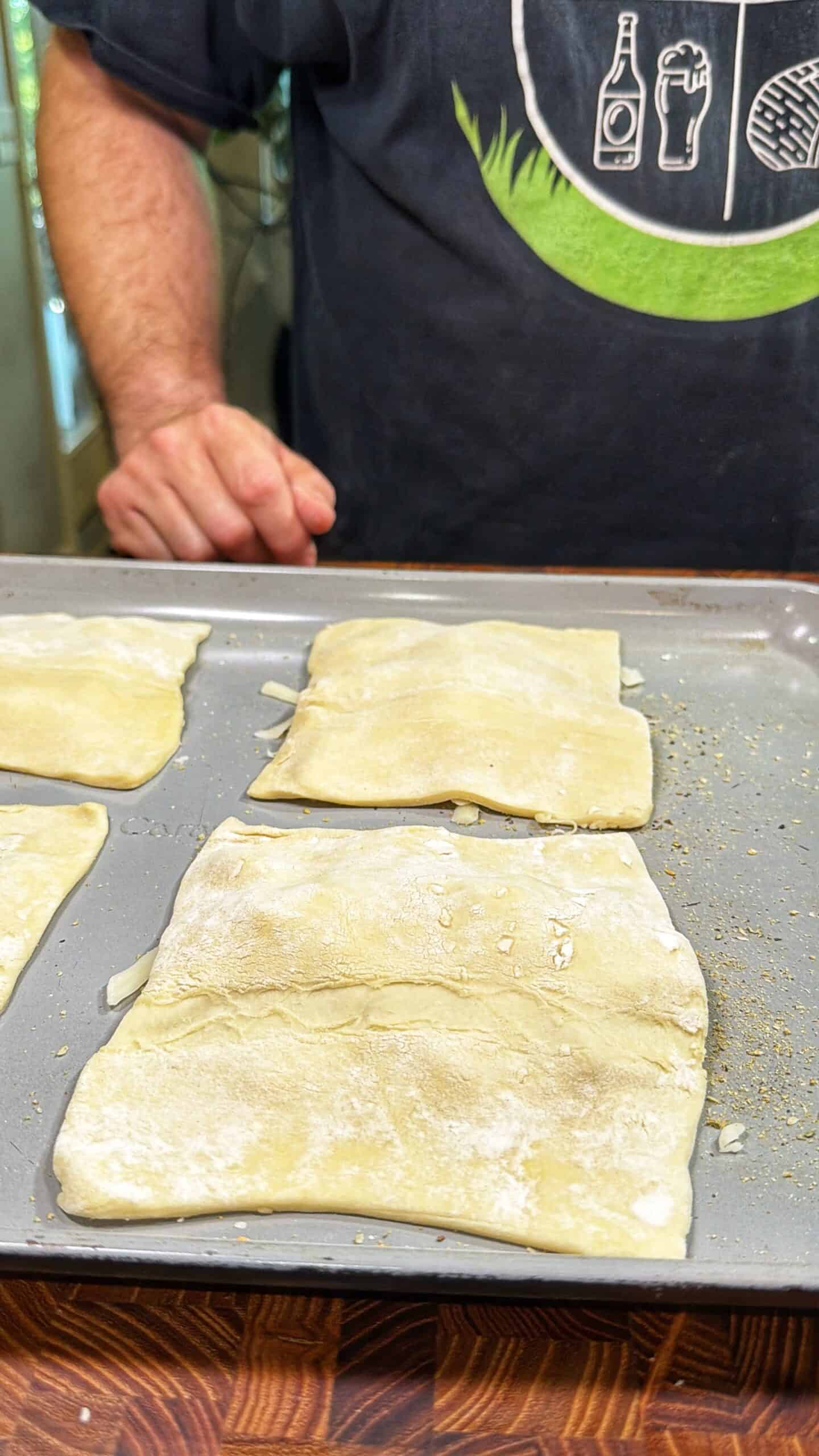 A person wearing a dark shirt stands behind a baking tray with three unbaked, square pieces of dough placed on it, ready to be baked.