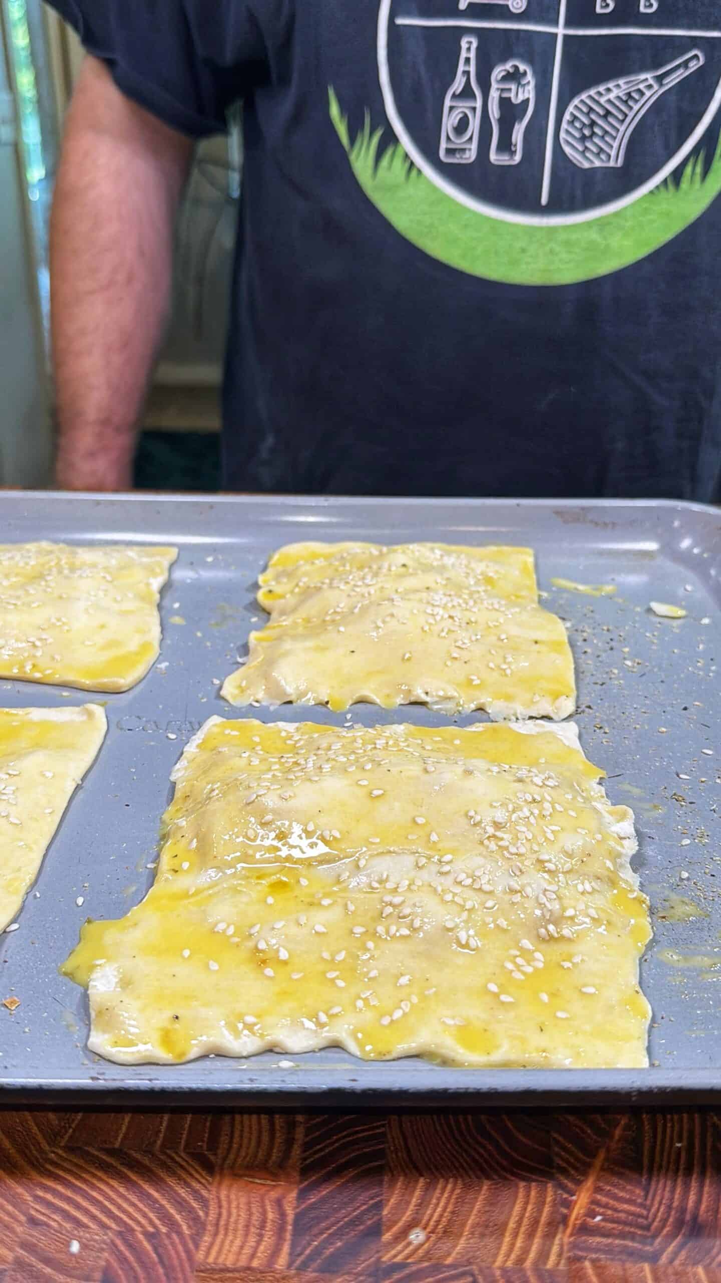 Rectangular pastry pieces brushed with egg wash and sprinkled with sesame seeds are on a baking tray. A person wearing a black shirt featuring various icons stands behind the tray.