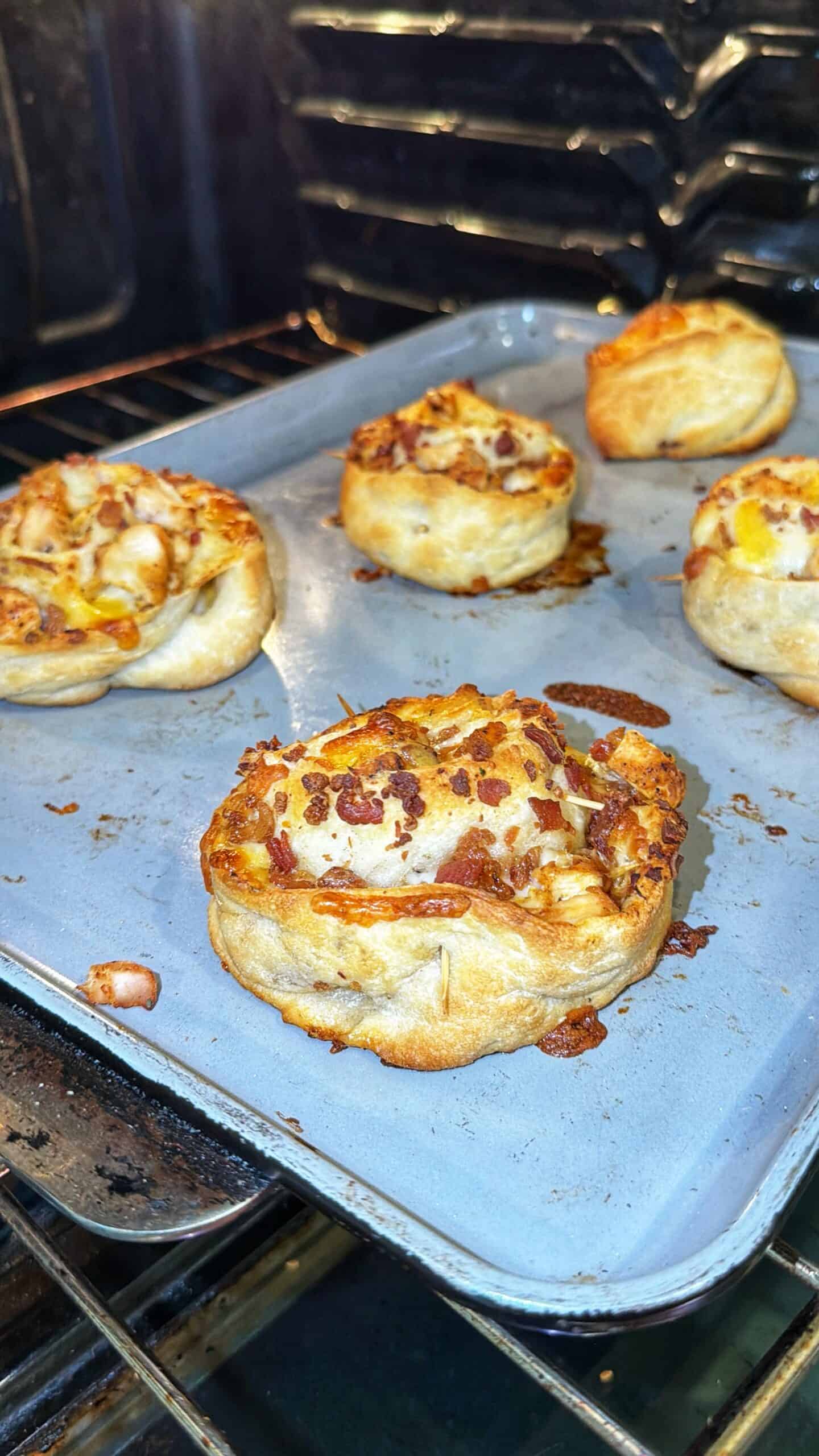 Five baked pizza rolls topped with cheese and bacon bits sit on a baking tray inside an oven. The rolls are golden brown with melted cheese and crispy toppings.