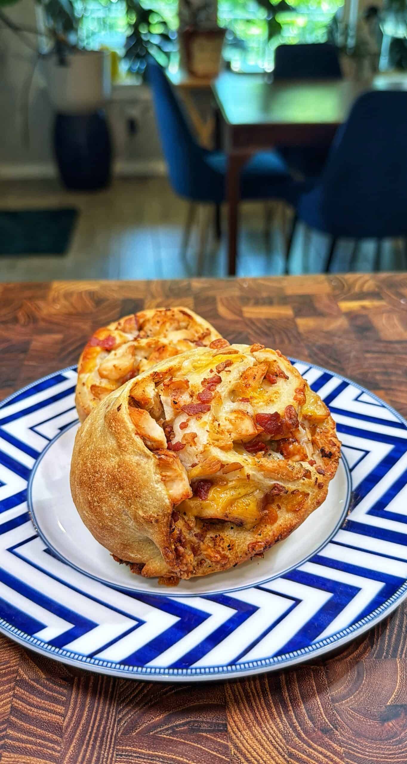 Two golden-brown, cheese and meat-filled bread rolls sit on a white plate with blue zigzag patterns, placed on a wooden table with a dining area and plants visible in the background.