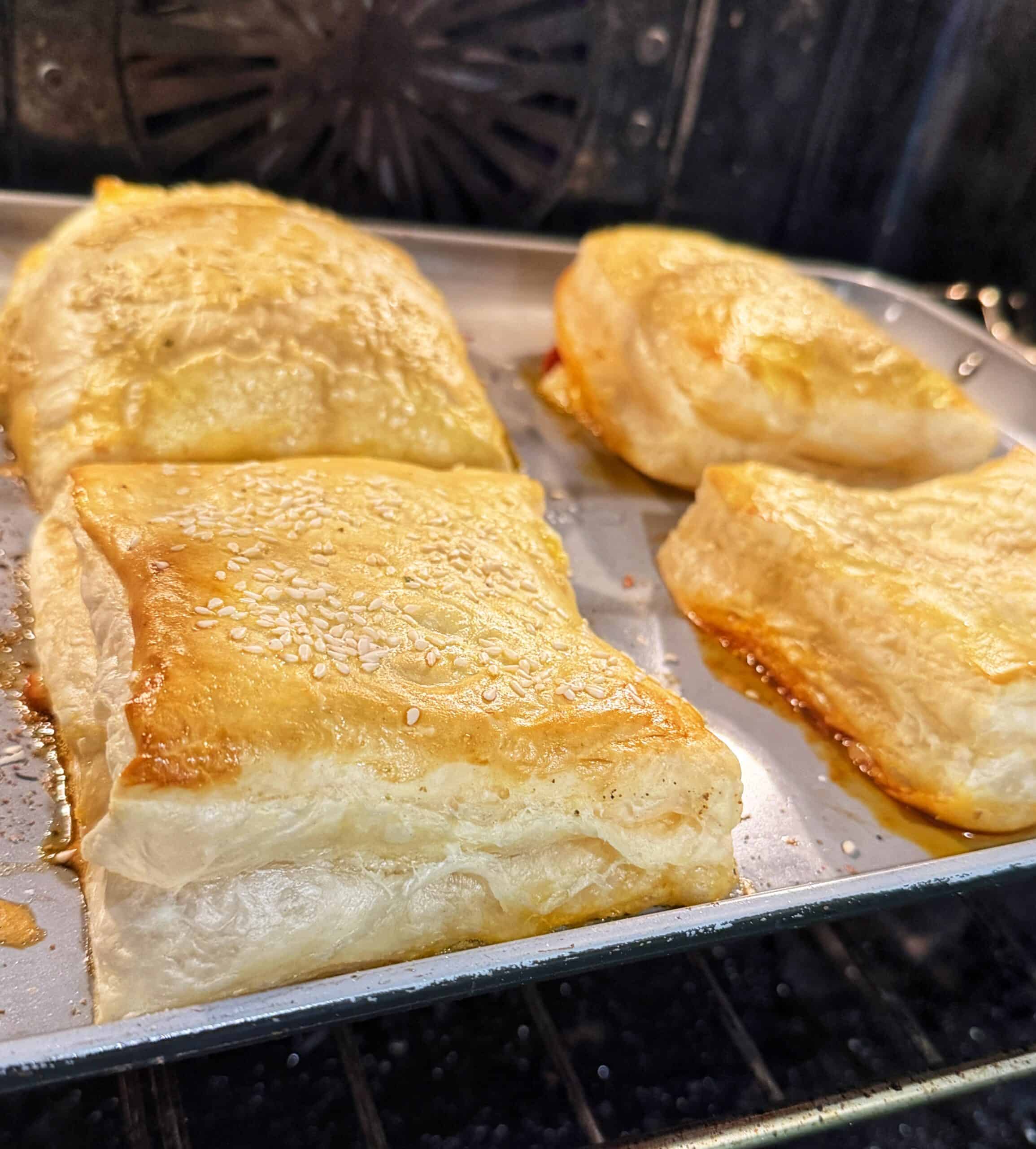 Four golden, flaky puff pastry squares sprinkled with sesame seeds are baking on a tray in an oven. Some filling is slightly oozing from the sides, and the pastries appear fresh and homemade.
