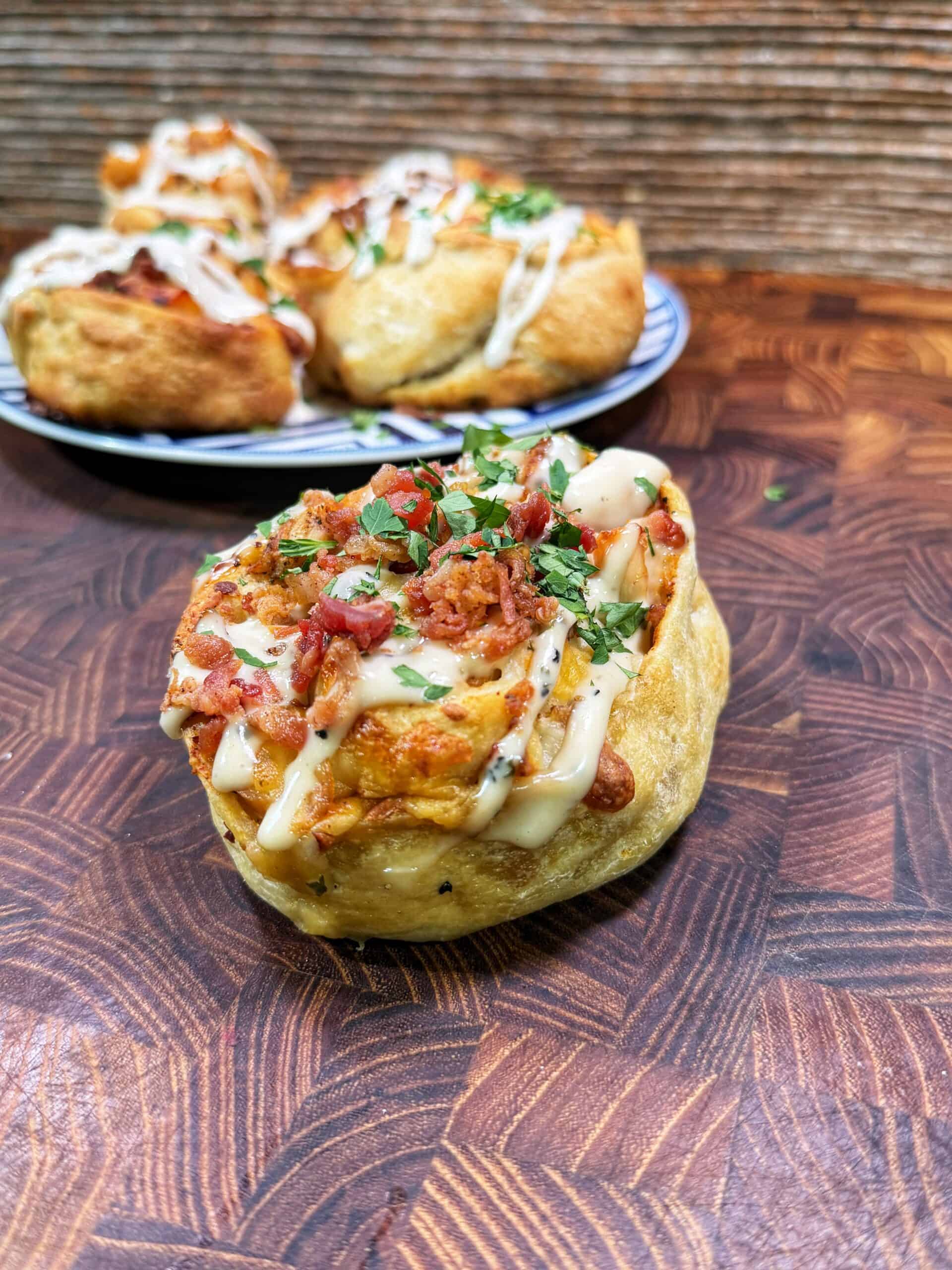 A savory pastry topped with chopped herbs, diced bacon, and drizzled with a creamy sauce sits on a wooden surface. In the background, more pastries are arranged on a plate.