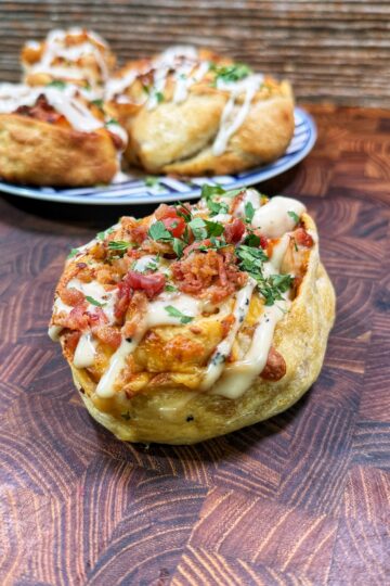 A close-up of a savory bread roll topped with bacon, herbs, melted cheese, and a drizzle of white sauce, placed on a wooden surface. More rolls are on a plate in the background.