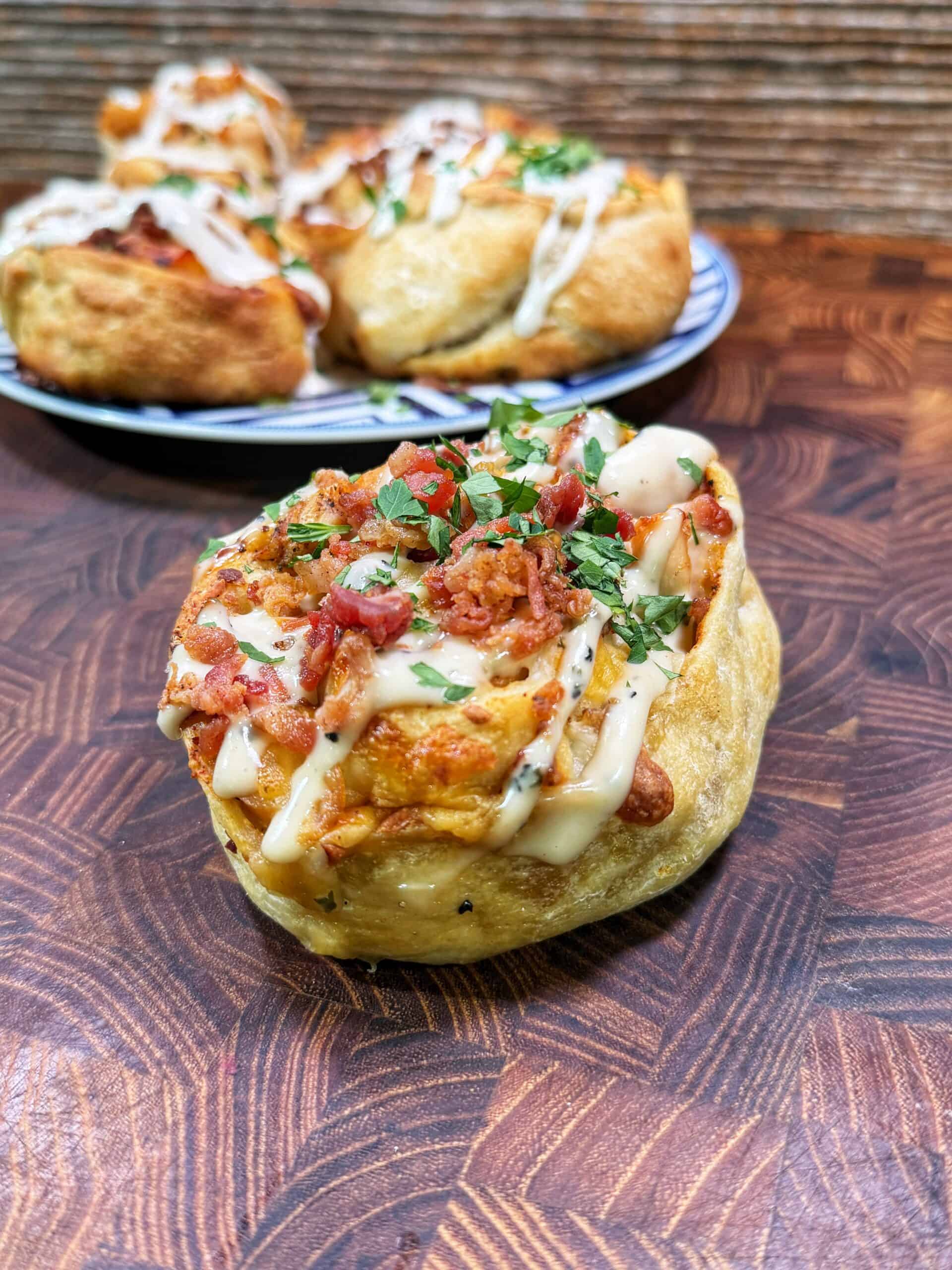 A close-up of a savory bread roll topped with bacon, herbs, melted cheese, and a drizzle of white sauce, placed on a wooden surface. More rolls are on a plate in the background.